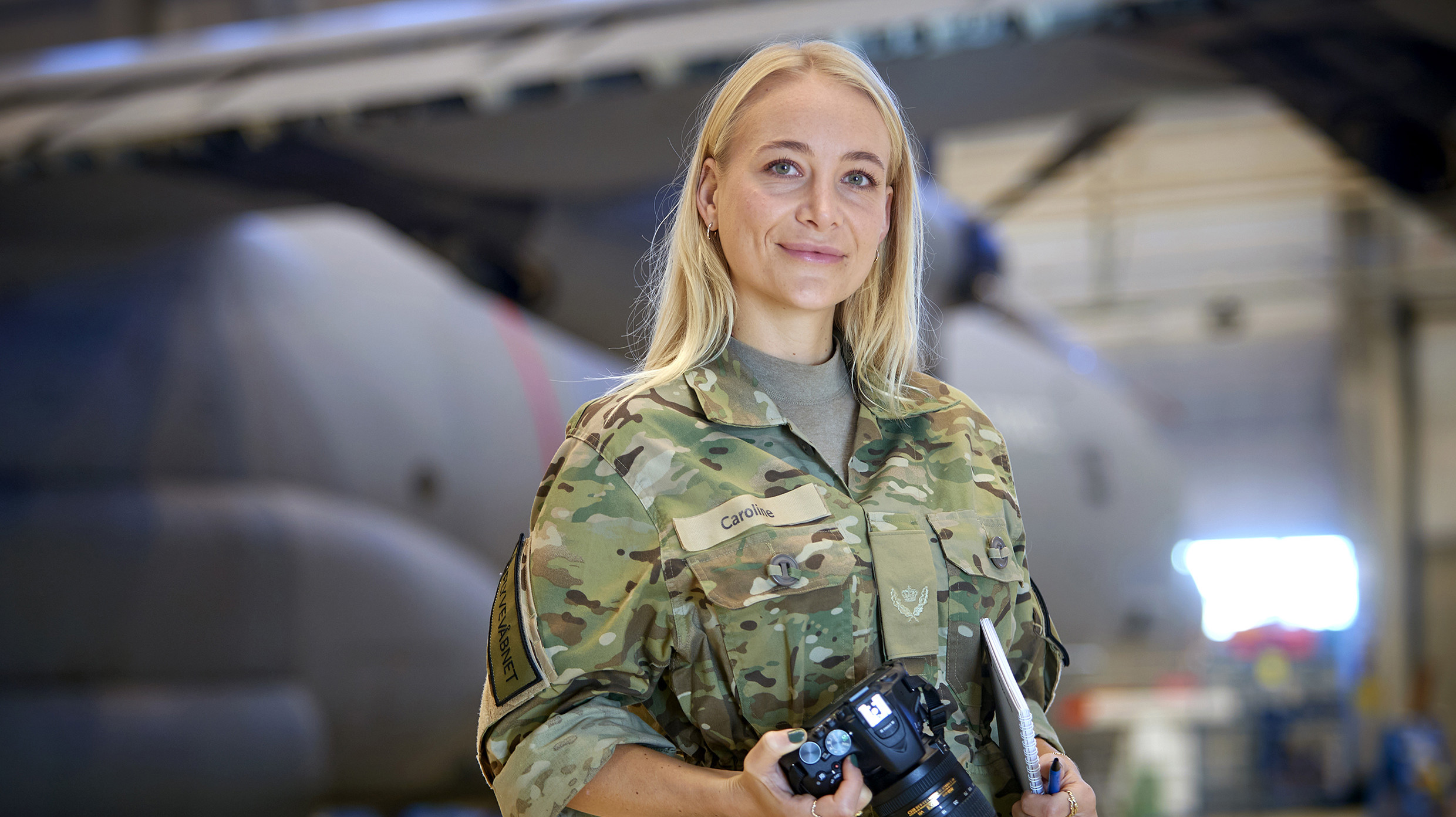 En kvinde iført militær camouflagetøj står selvsikkert i en flyhangar med et kamera i den ene hånd og en notesbog i den anden. I baggrunden ses silhuetten af en helikopter, hvilket understreger det militære miljø. Hendes navneskilt viser "Caroline". Situationen udstråler professionalisme og engagement, sandsynligvis i forbindelse med dokumentation eller mediearbejde inden for militæret.
