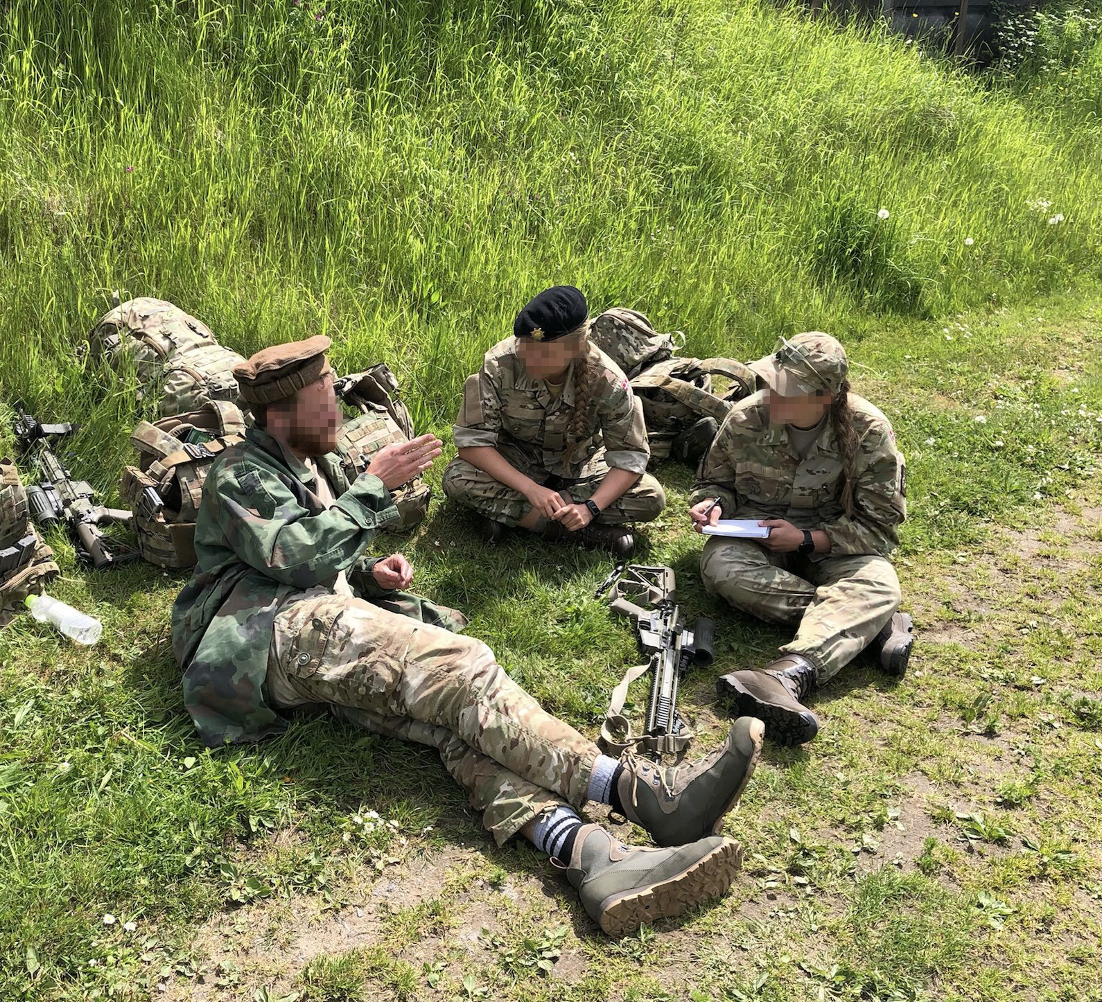 Militærpersonale sidder på græsset i samtale, hvor én forklarer og de andre lytter; udstyr er spredt omkring dem.