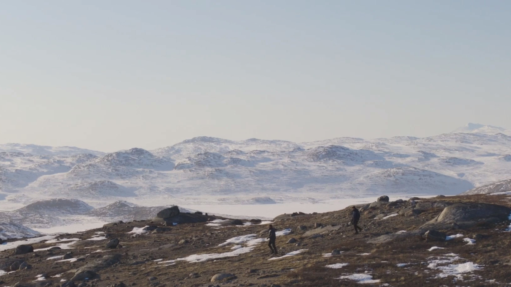 Sneklædt landskab med bakker og bjerge i baggrunden, to personer går gennem terrænet under en klar himmel.