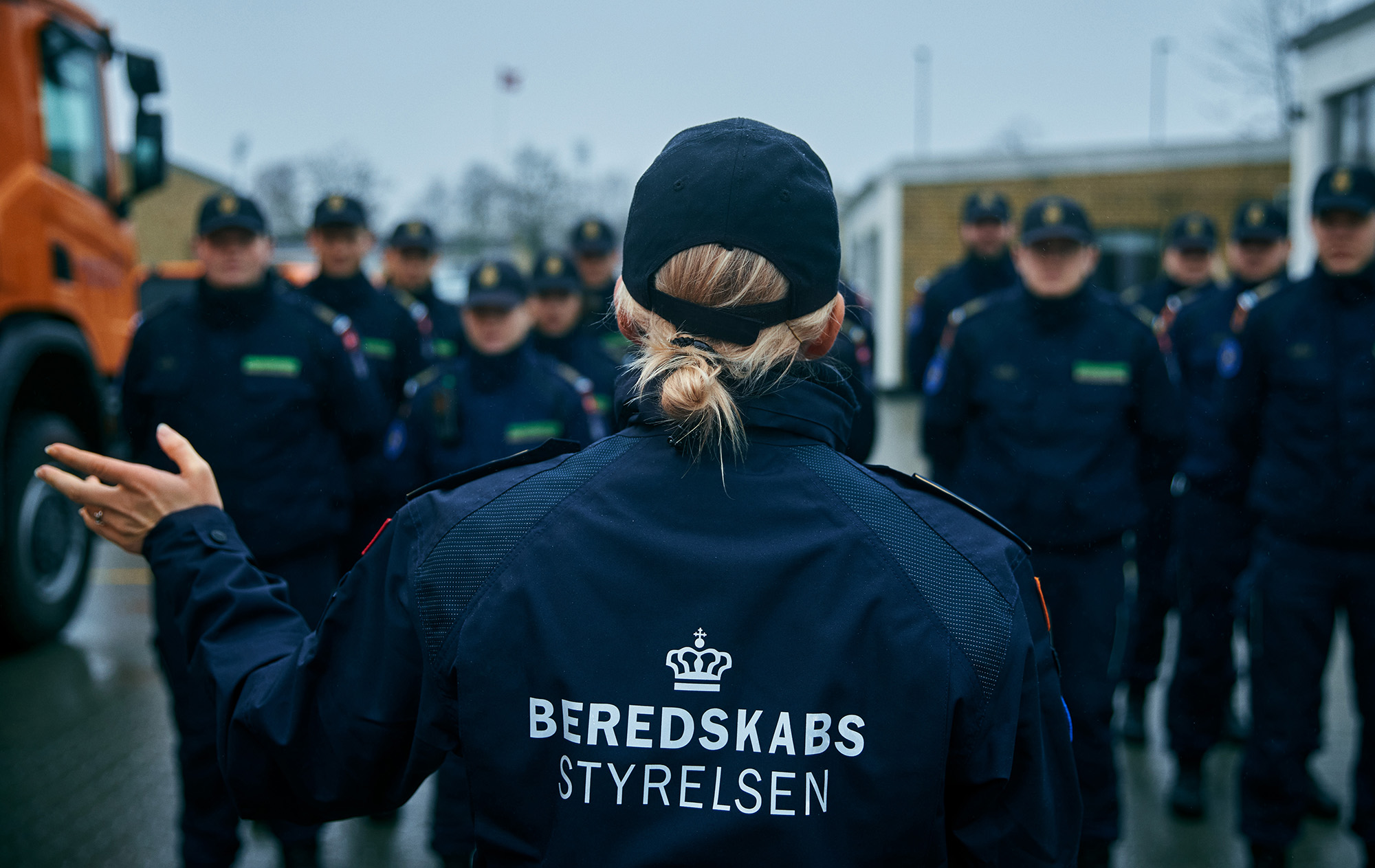 Person i uniform med hestehale står med ryggen til og giver briefing til gruppe i uniform ved industrielt anlæg under overskyet himmel.