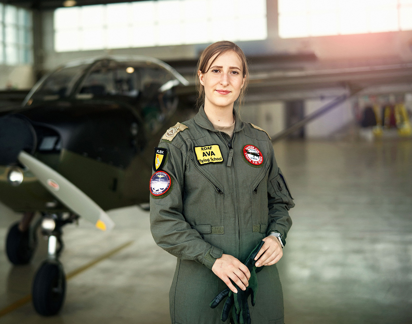 Kvinde i militærflyverdragt står selvsikkert foran et lille RDAF-fly i en hangar med store vinduer og naturligt lys.
