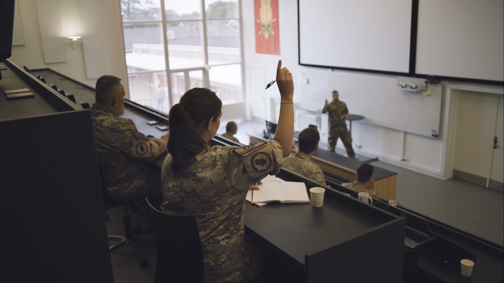 Kvinde i camouflageuniform rækker hånden op i et moderne klasseværelse med militærundervisning og stor projektorskærm.