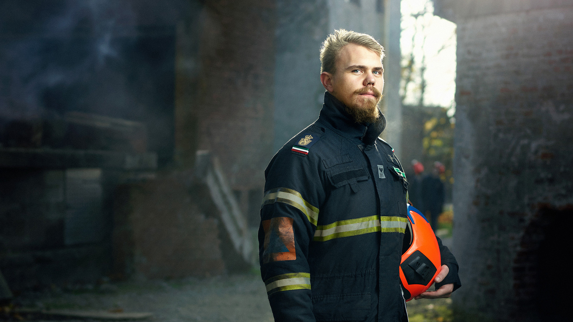 Brandmand med let skæg i mørk uniform med reflekser holder en orange hjelm, står seriøst i et industrimiljø med røg.
