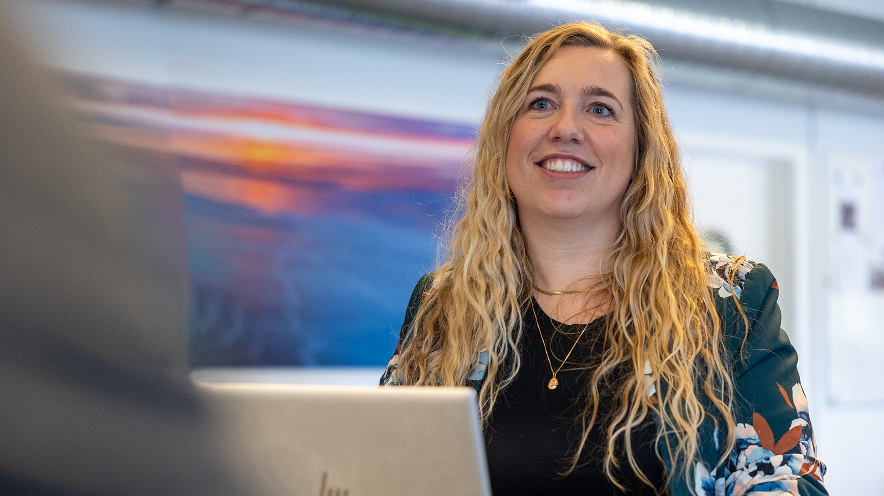 Kvinde med langt, bølget blondt hår og venligt smil sidder ved et bord med en bærbar computer, iført sort top og blomstret blazer. Farverige abstrakte kunstværker ses i baggrunden.