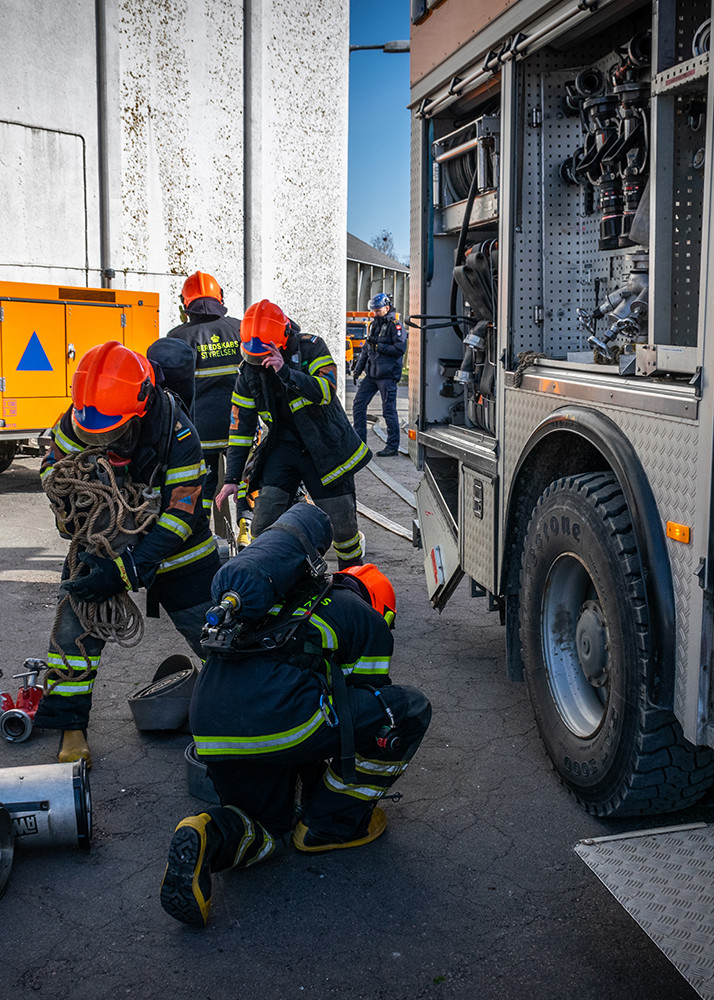Brandmænd i sorte uniformer med reflekser og orange hjelme arbejder sammen med brandslanger foran en brandbil.