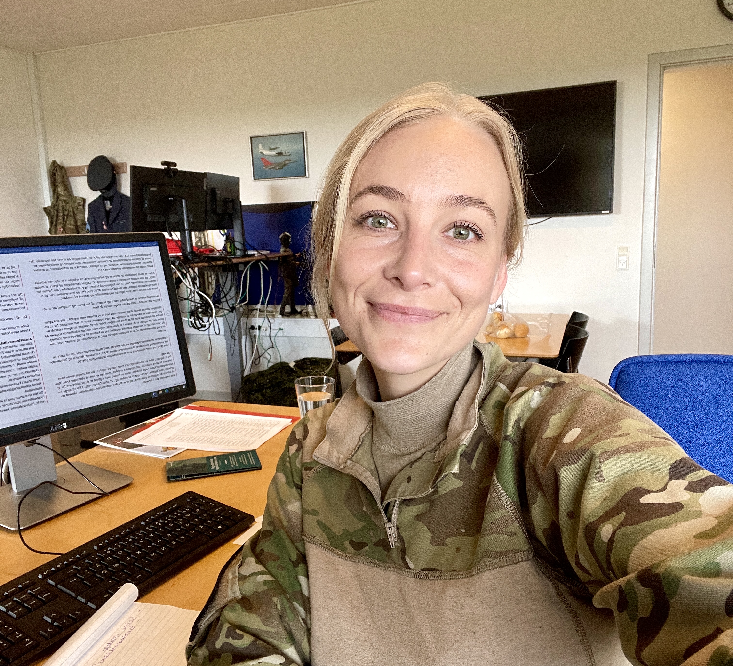Hovedpersonen på billedet er en smilende kvinde iført en militærjakke med camouflage-mønster. Hun sidder ved et skrivebord med en computerskærm, hvor der vises tekst, og der ligger forskellige genstande på bordet, herunder papirer og et glas vand. I baggrunden ses flere skærme og nogle udstillede militæruniformer. Omgivelserne ligner et arbejdsmiljø eller kontor med tilknytning til militær- eller forsvarsopgaver.