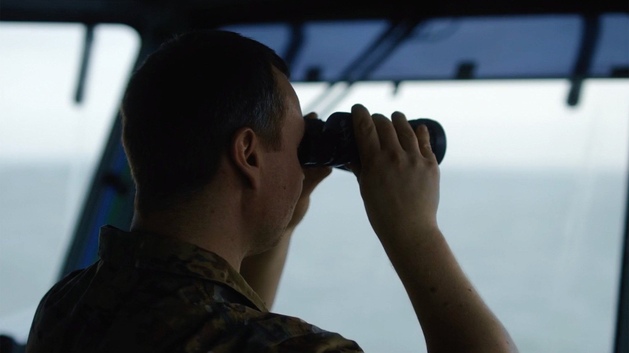 En person i camouflageuniform står på et skibs bro og spejder ud mod horisonten med en kikkert, mens de er intenst fokuseret på havudsigten i det svagt oplyste interiør.