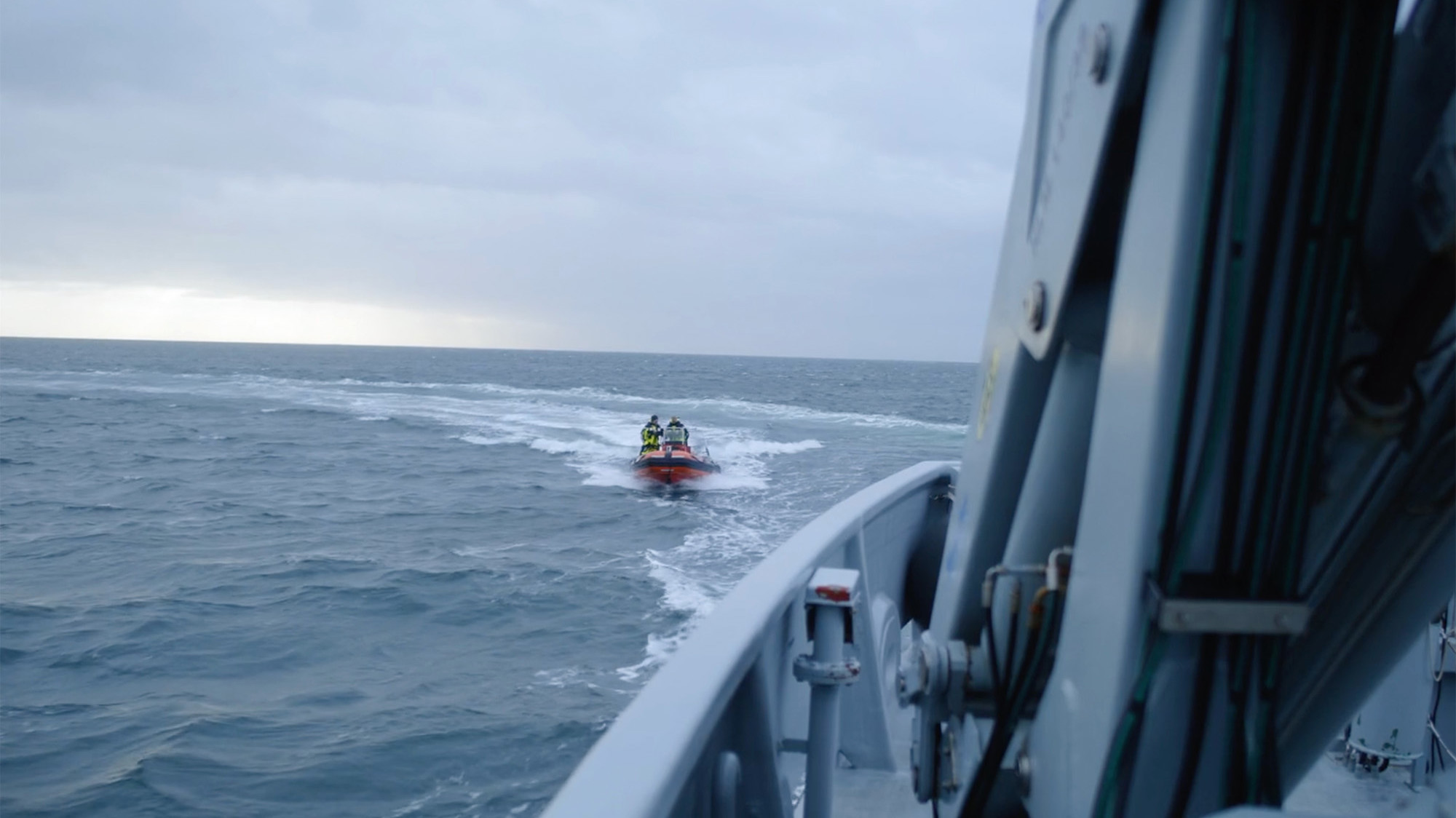 Billedet viser en kystvagts- eller redningsaktion til søs. Hovedmotivet er en lille orange redningsbåd i det fjerne, der sejler gennem bølgerne. To personer i sikkerhedsudstyr er synlige på båden, hvilket antyder en igangværende mission. I forgrunden ses kanten af et større fartøj, som giver kontekst til operationen. Himlen er overskyet, hvilket indikerer potentielt udfordrende vejrforhold.