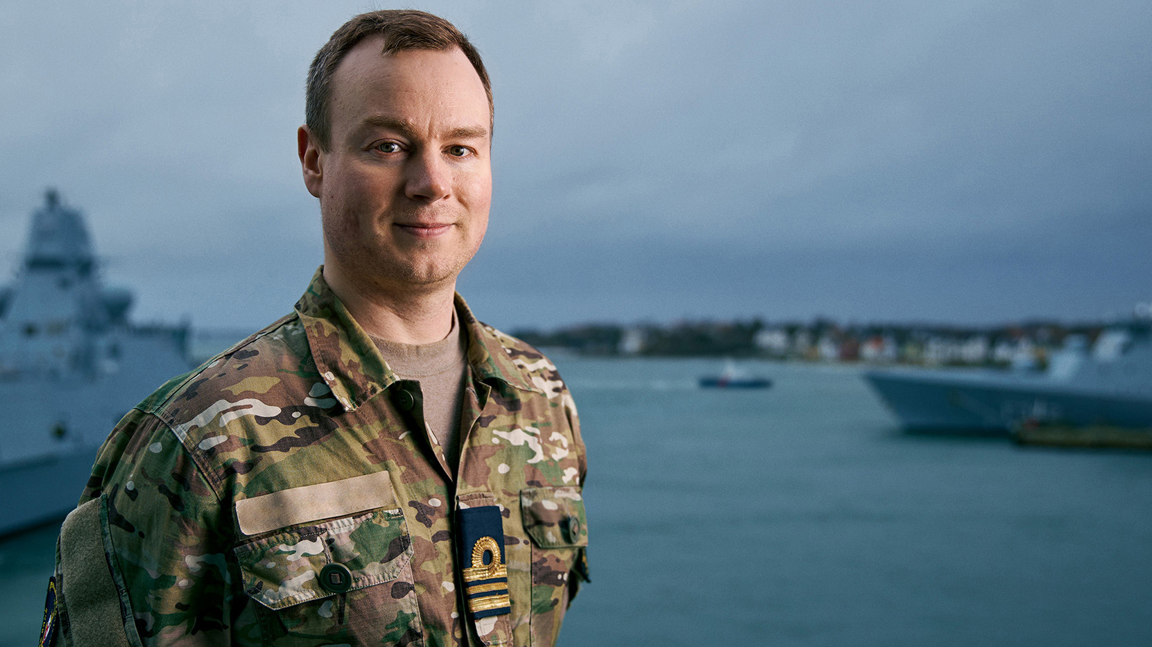 En mand i militærkamuflageuniform smiler og står foran et vandområde med skibe i baggrunden. Han har et militærmærke eller en ranginsignie på uniformen, hvilket indikerer rang eller position. Omgivelserne ligner en flådemiljø med overskyet himmel og roligt farvand, hvilket antyder en maritim operation eller base.