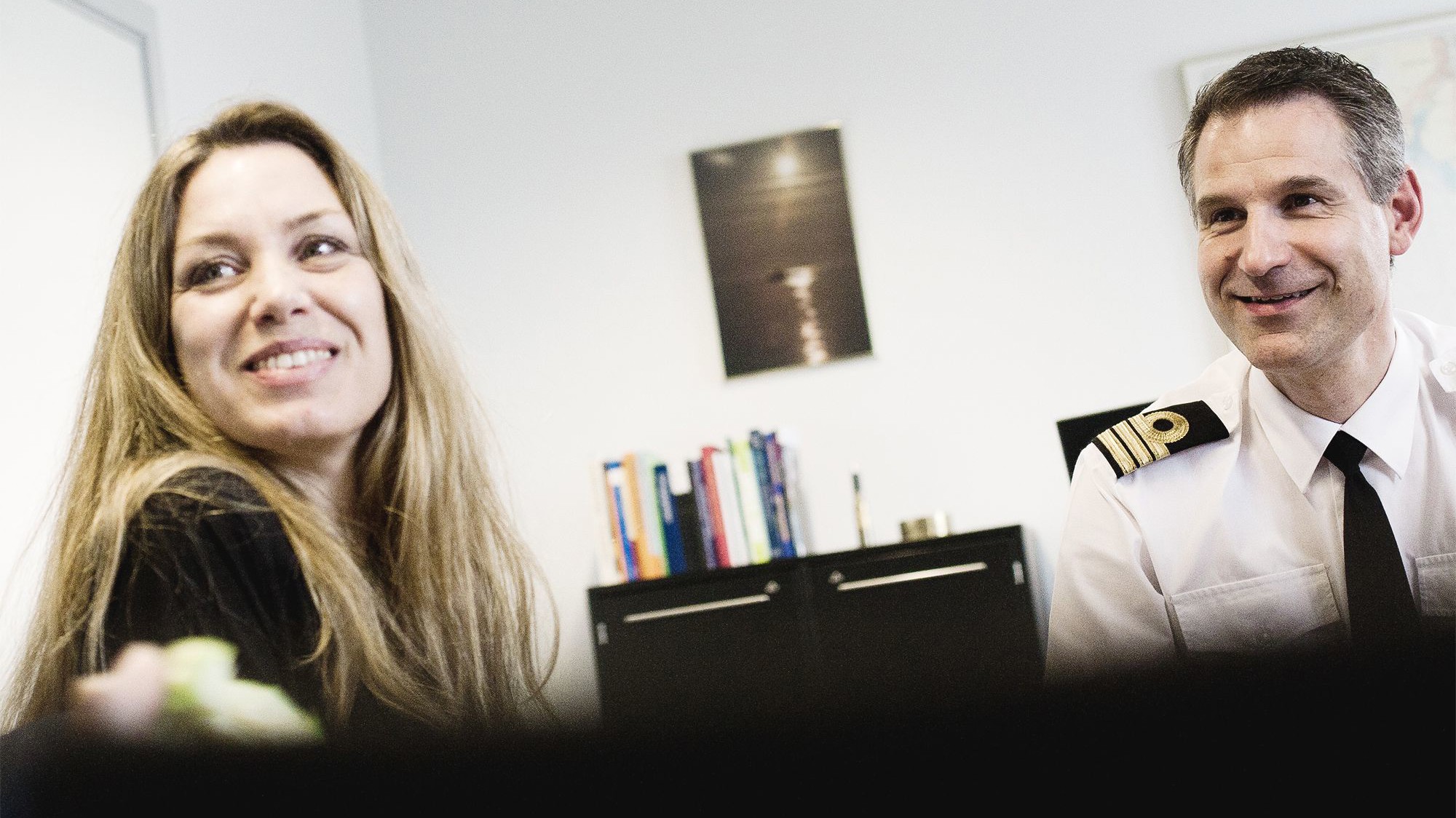 Kvinde med langt, bølget hår og mand i uniformsskjorte med skuldermærker smiler i et afslappet, professionelt kontormiljø.