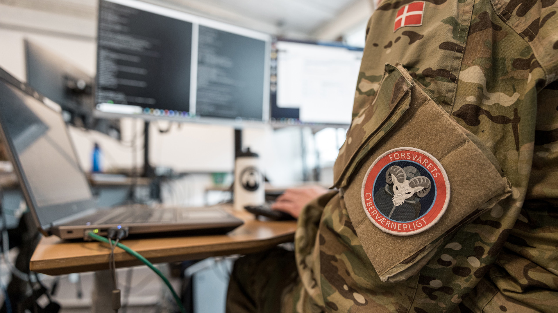 Person i dansk camouflageuniform med mærke "FORSVARETS CYBERVÆRNEPLIGT" ved et skrivebord med to skærme og en bærbar computer.