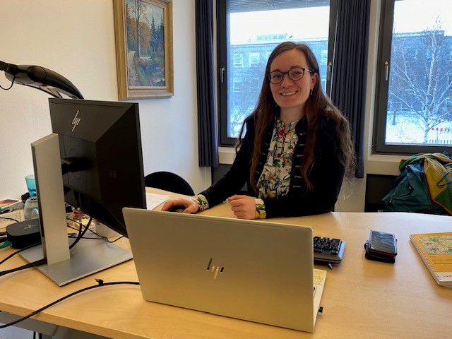 Person med langt hår og briller smiler ved et skrivebord i et kontormiljø med to skærme og udsigt til et snedækket landskab.