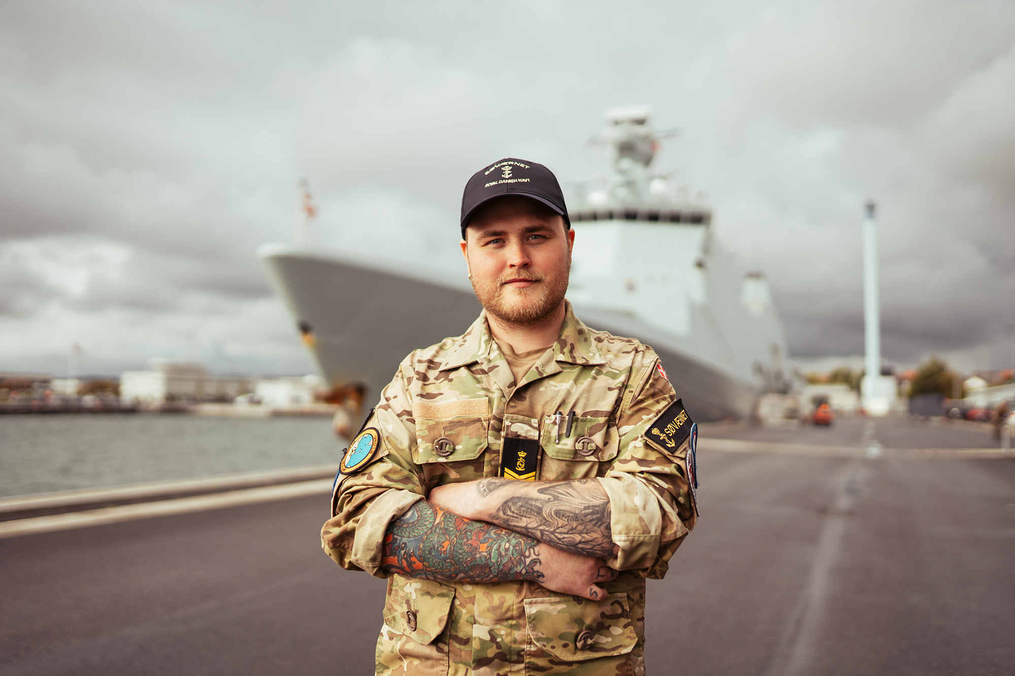 Militærperson i camouflagedragt med korslagte arme og synlige tatoveringer, står foran et krigsskib i havn under skyet himmel.