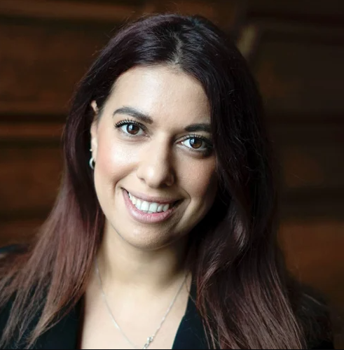 Billedet viser en kvinde med langt, mørkt hår med rødlige reflekser. Hun smiler og kigger direkte ind i kameraet, hvilket giver et venligt og imødekommende udtryk. Hun bærer øreringe og et halskæde, og baggrunden antyder en varm, træbeklædt atmosfære. Hendes tøj fremstår professionelt og understreger hendes velplejede udseende.