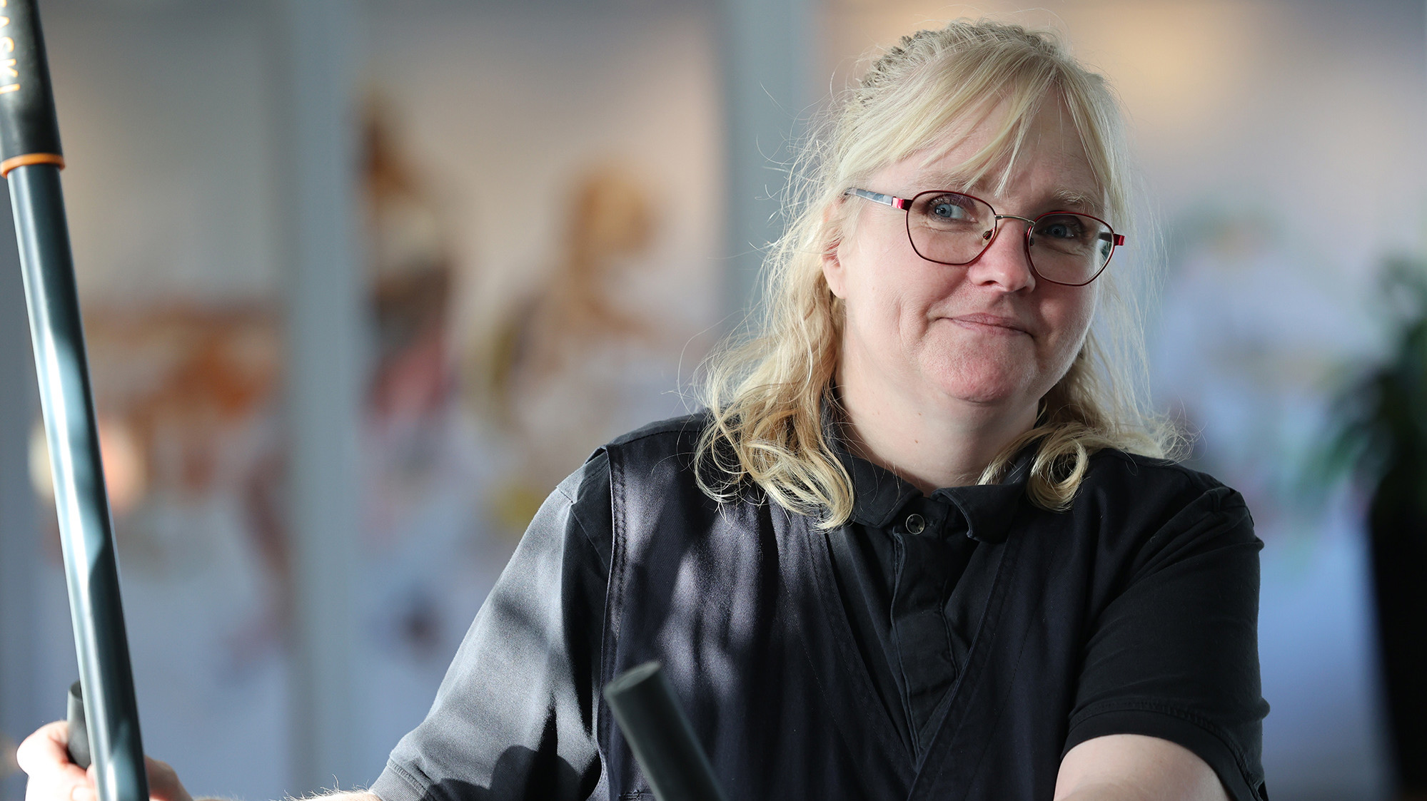 Billedet viser en kvinde med blondt hår, briller og en mørk skjorte. Hun holder et rengøringsredskab og smiler eller ser engageret ud, hvilket antyder et venligt udtryk. Baggrunden har bløde, slørede farver og former, hvilket indikerer et lyst, måske kunstnerisk miljø. Stemningen er afslappet og imødekommende.