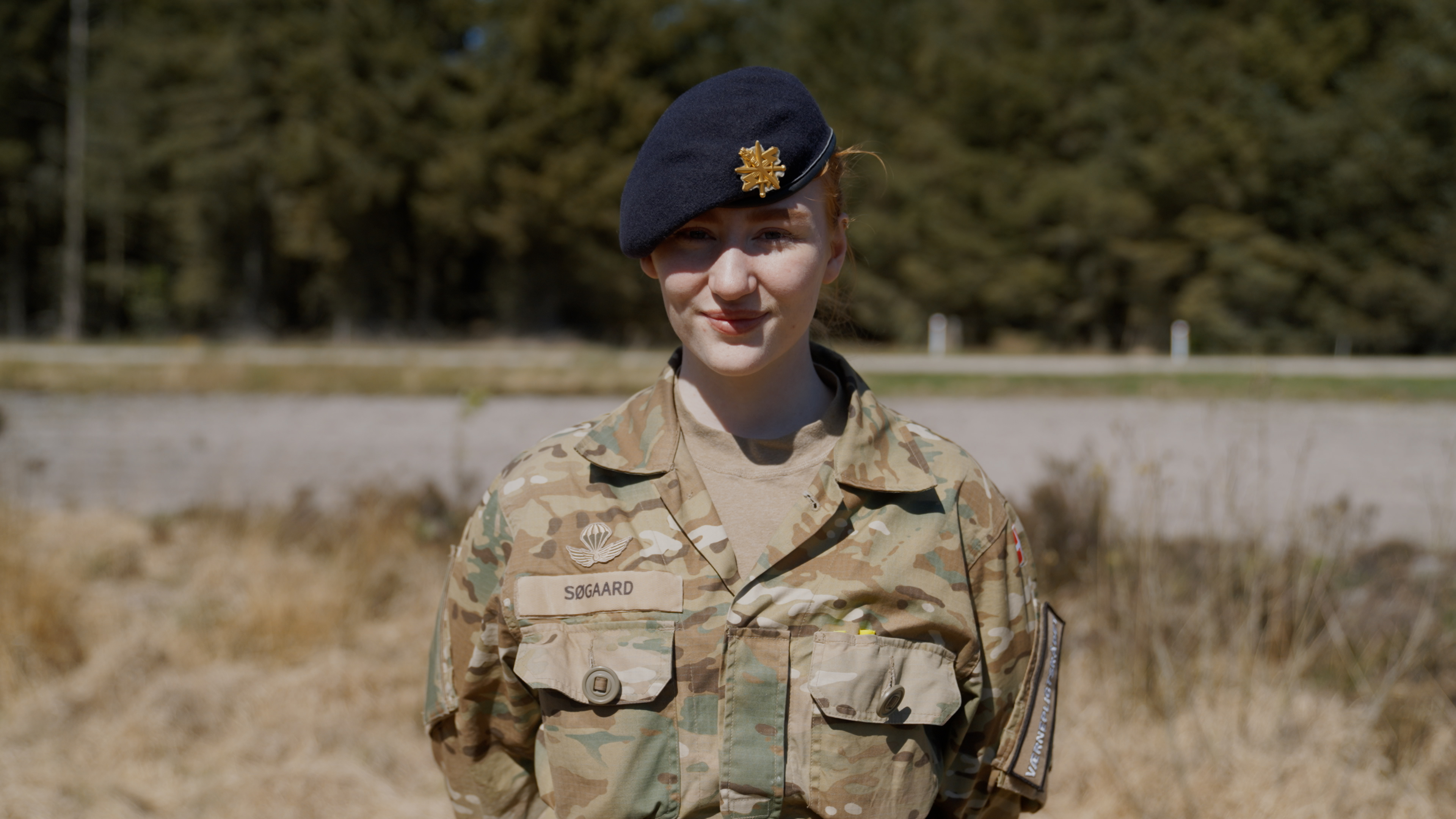 Militærperson i camouflageuniform og mørk baret med insignie, navneskiltet siger "Søgaard", udendørs omgivet af natur.