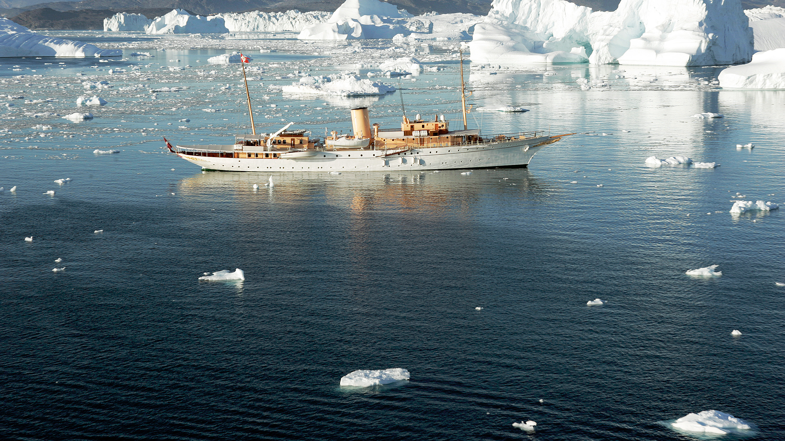 Klassisk yacht med trædetaljer og to master sejler mellem isbjerge i klart, spejlende vand i et roligt, polart landskab.