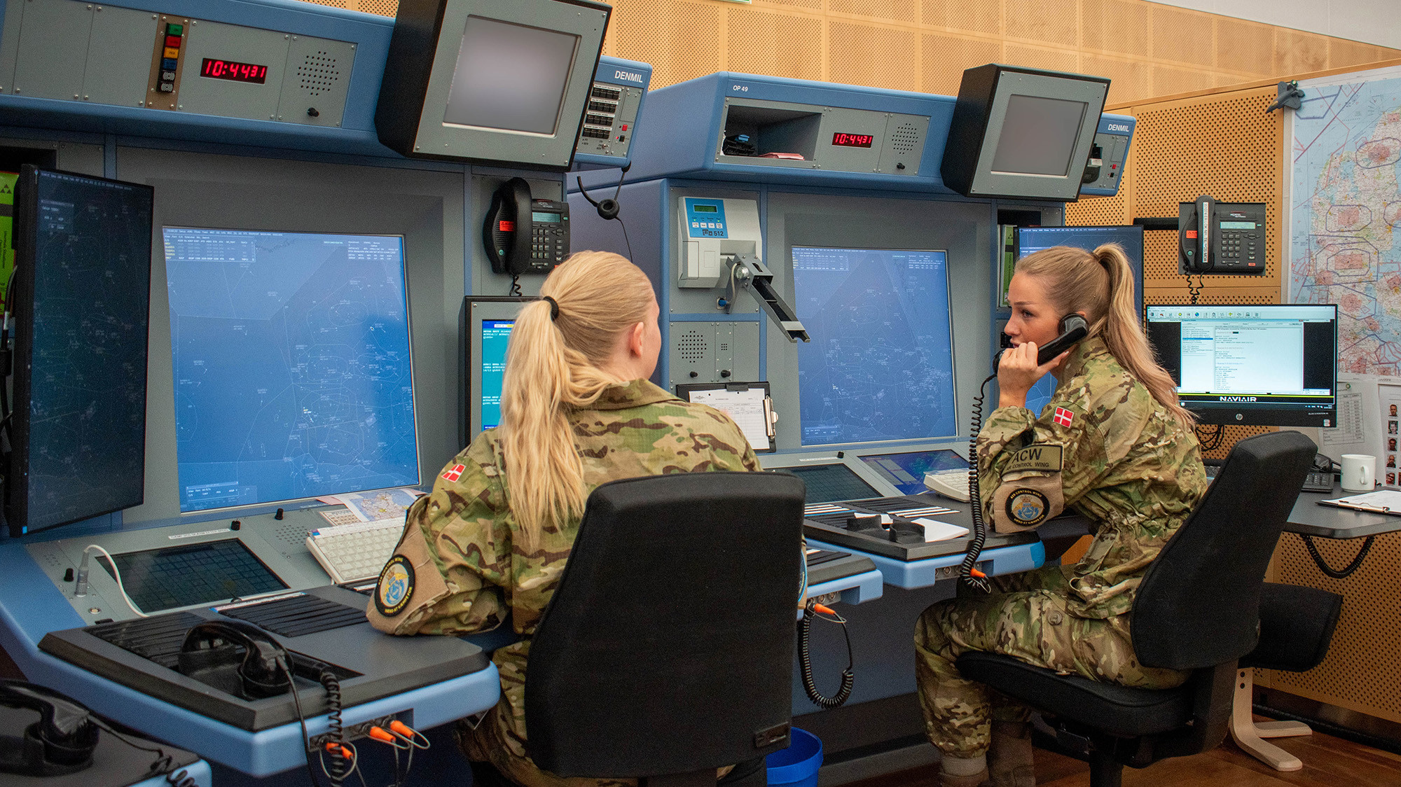 To militærpersoner i camouflageuniform arbejder ved skriveborde med flere computerskærme og radar i et kontrolrum med avanceret udstyr.