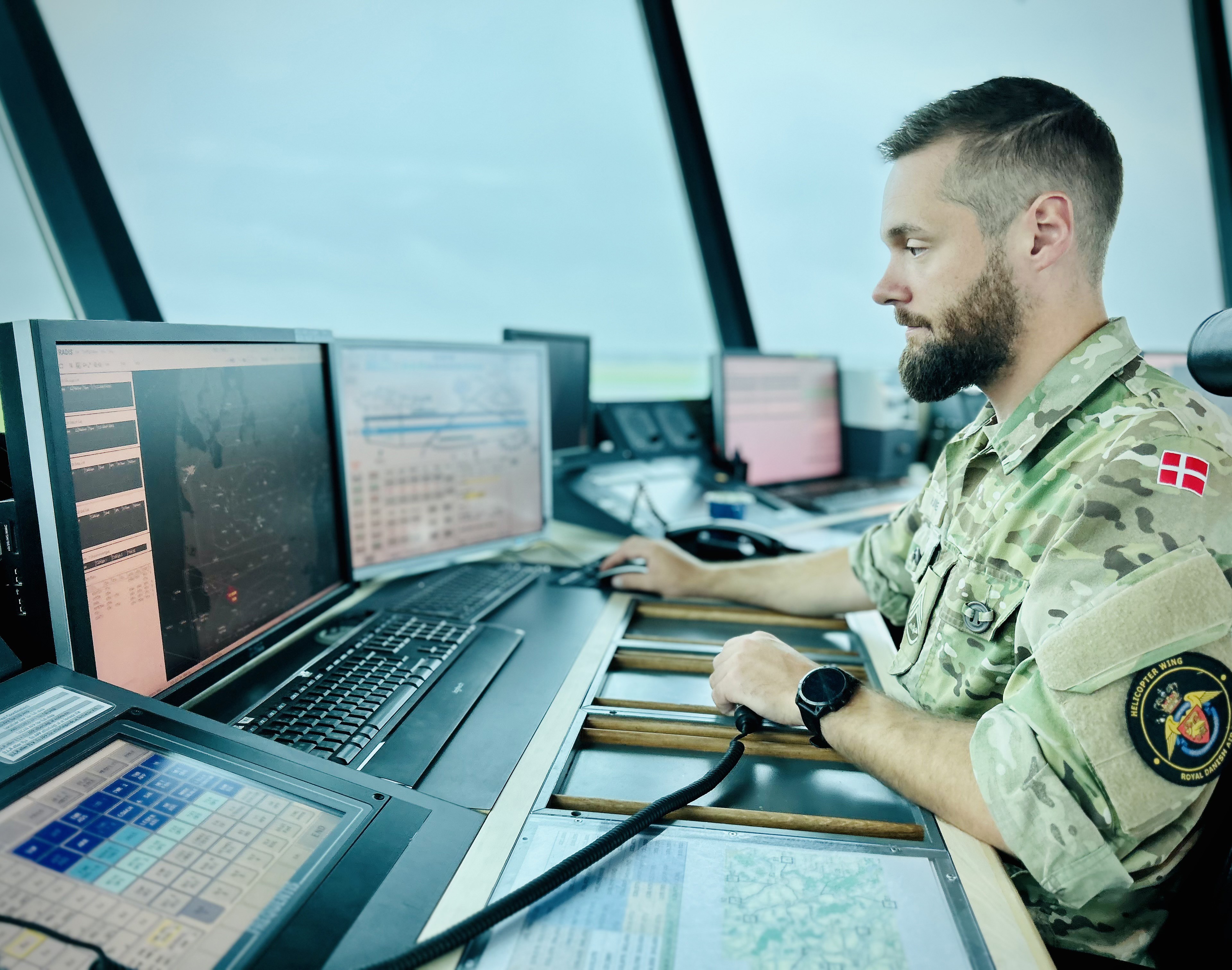 Dansk militærperson i camouflageuniform ved kontrolstation med danske flag og badge, omgivet af skærme og udsigt gennem store vinduer.