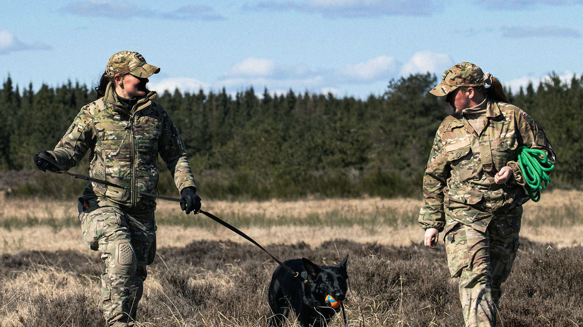 To personer i camouflageuniformer går med en sort hund med orange bold i munden i et græsbevokset, udendørs område.