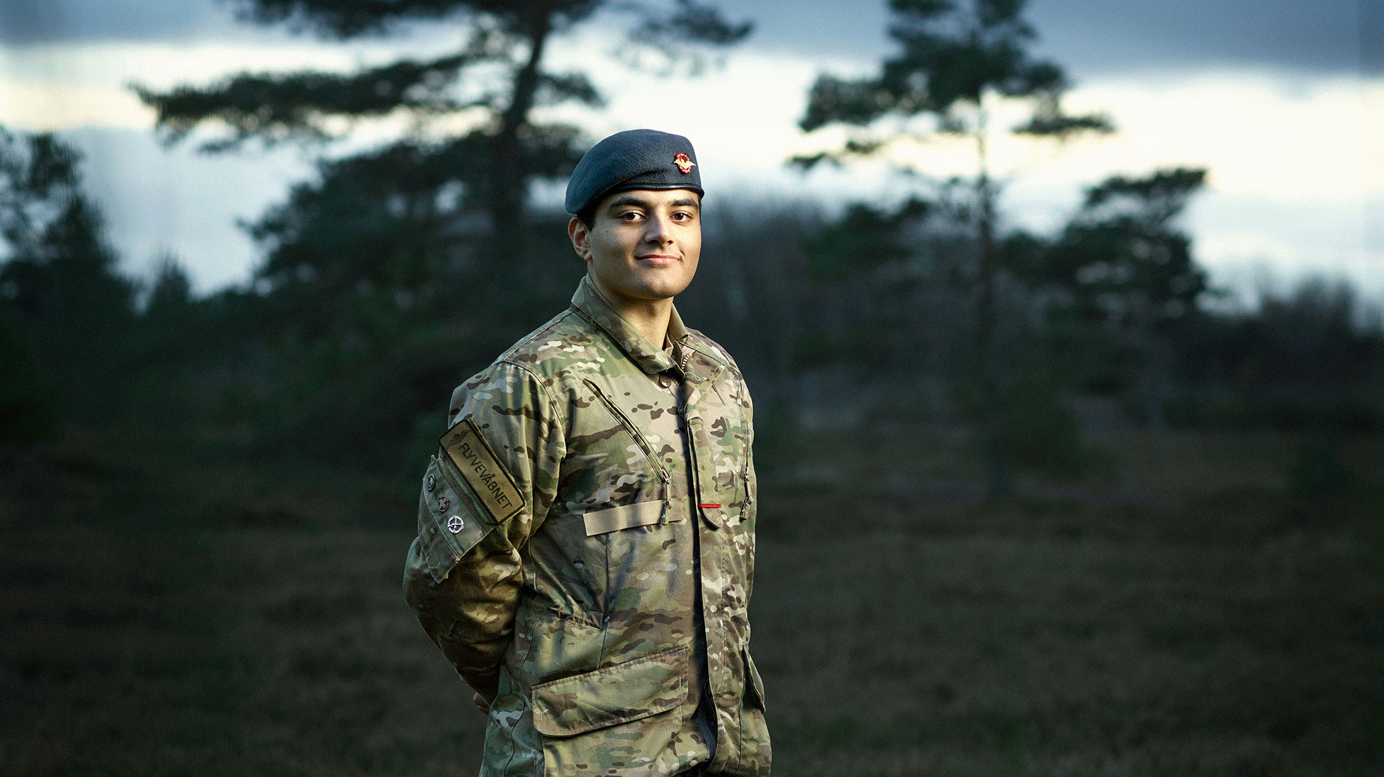 Ung person i militæruniform står selvsikkert blandt træer, med kasket og mærke på ærmet, ansigtet oplyst med dramatisk stemning.