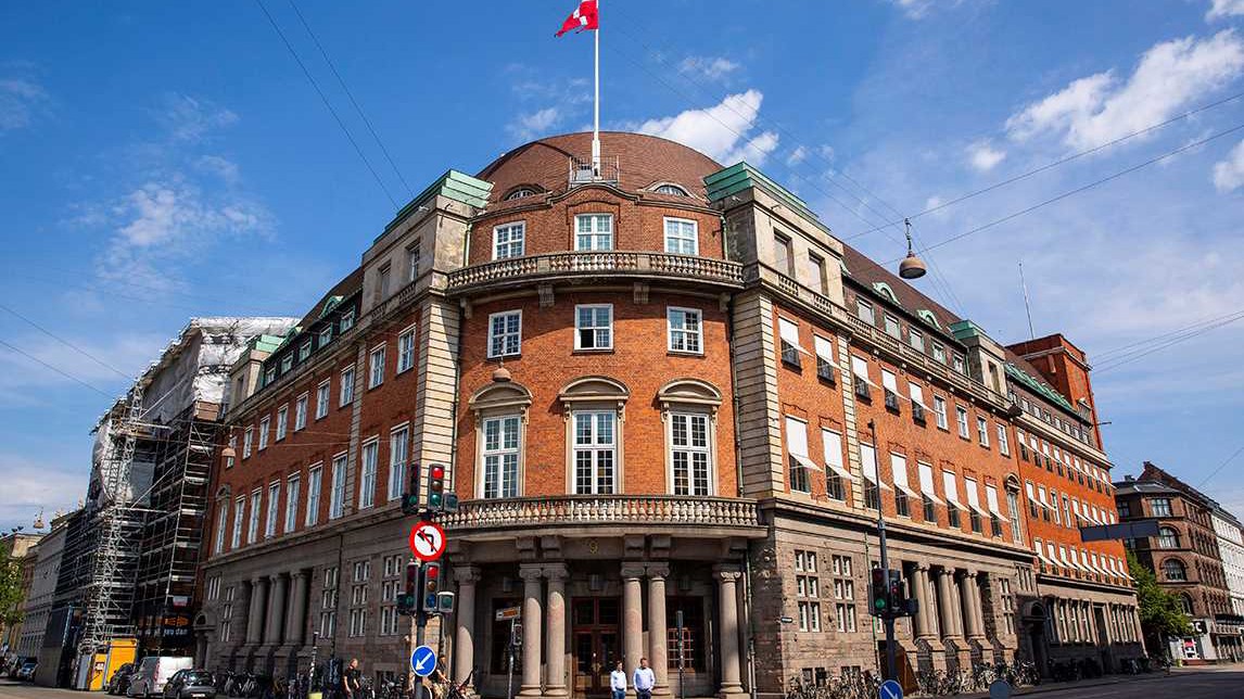 Et markant murstensbygning med klassisk arkitektur, buet tag og flere vinduer. Detaljer inkluderer et schweizisk flag, der vajer over bygningen, samt flere trafiklys foran, hvilket antyder et bymiljø. Facaden har store vinduer og detaljeret stenhuggerarbejde, hvilket tyder på historisk betydning. Til venstre ses en delvist synlig byggeplads, som indikerer igangværende renoveringer eller udvikling i nærheden.