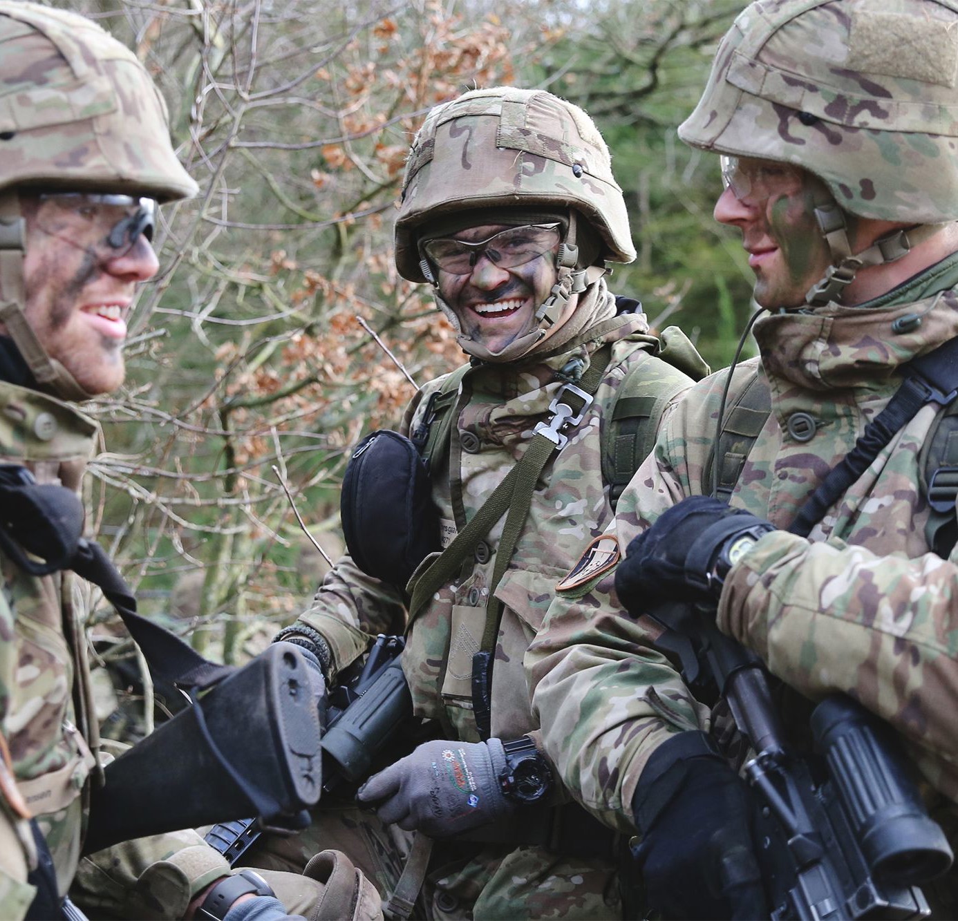 Tre værnepligtige soldater i camouflageuniform og hjelm smiler og taler sammen under en øvelse i skoven. De bærer udstyr og våben, og stemningen er afslappet og kammeratlig.