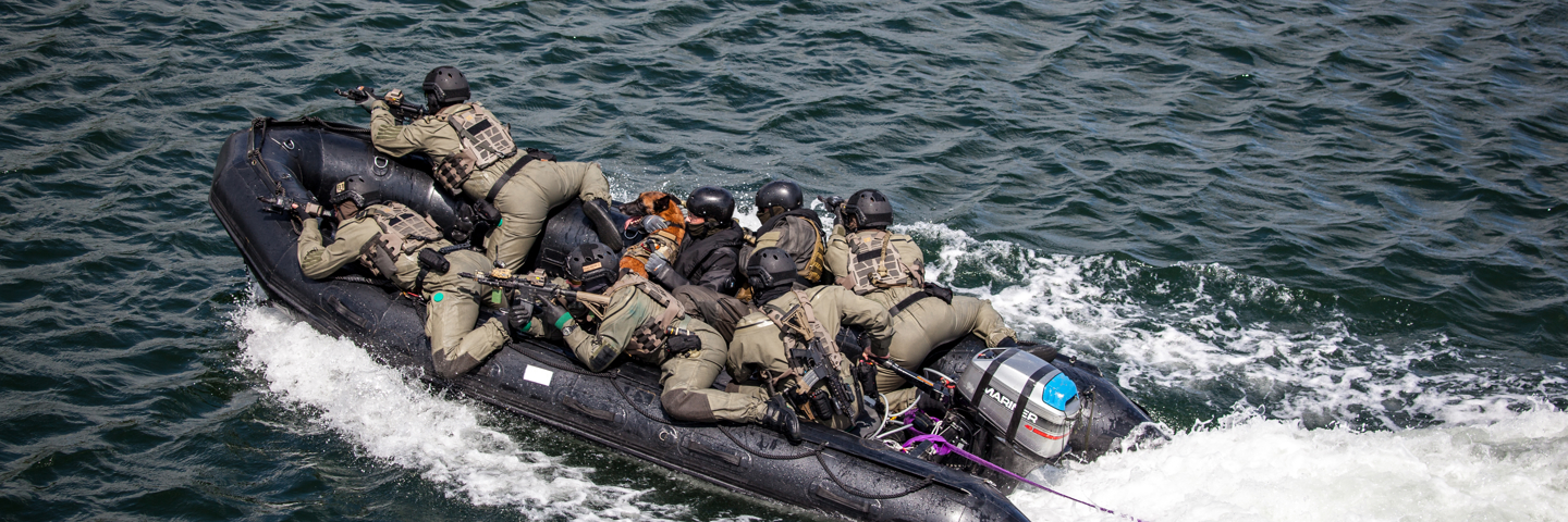 En gruppe personer i taktisk udstyr på en sort gummibåd bevæger sig hurtigt gennem vandet, koncentrerede og målrettede.