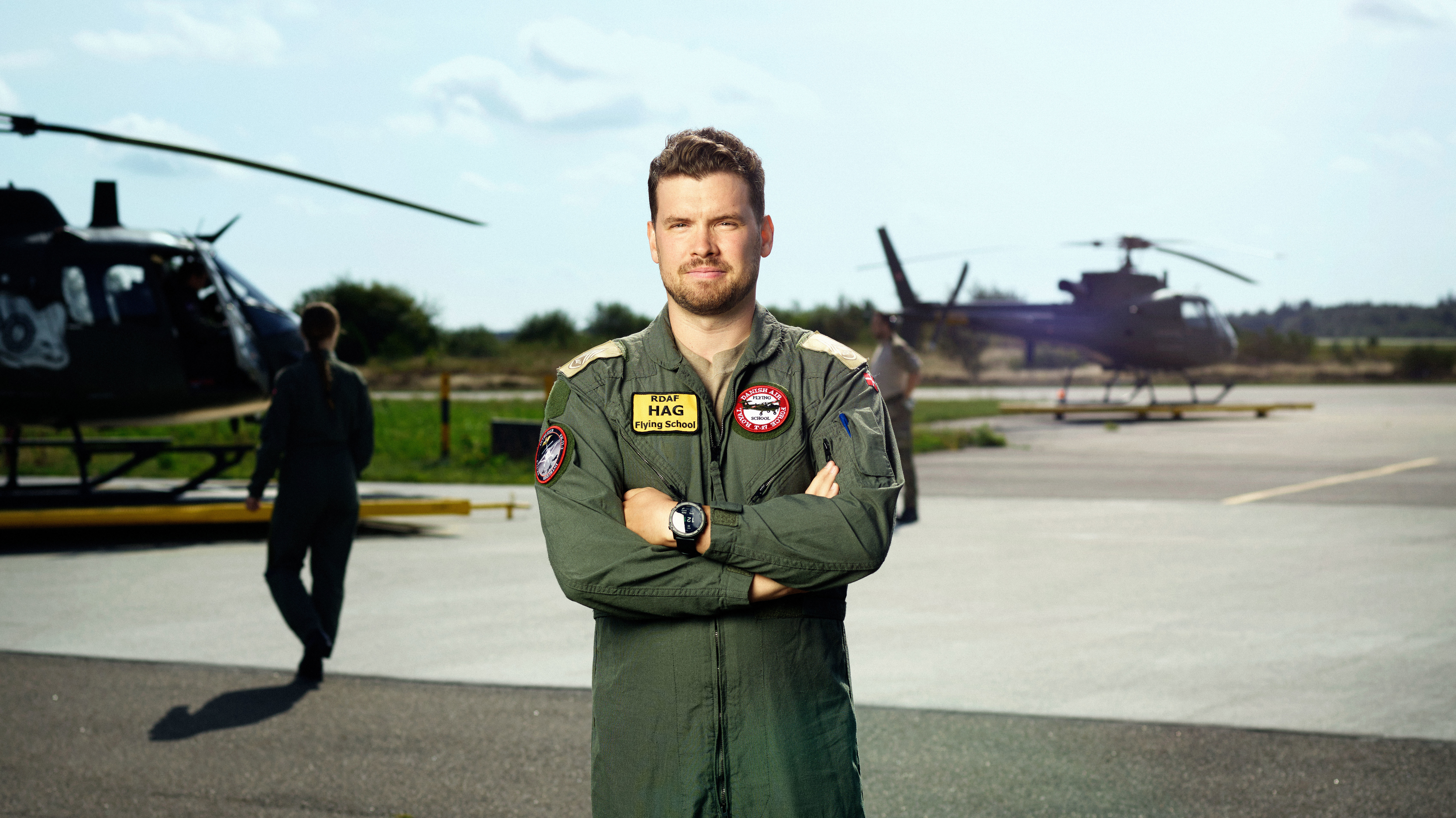 Mandlig helikopterpilot i grøn flyverdragt med RDaf Flying School-emblemer står selvsikkert på en solrig landingsplads med to helikoptere bag sig.