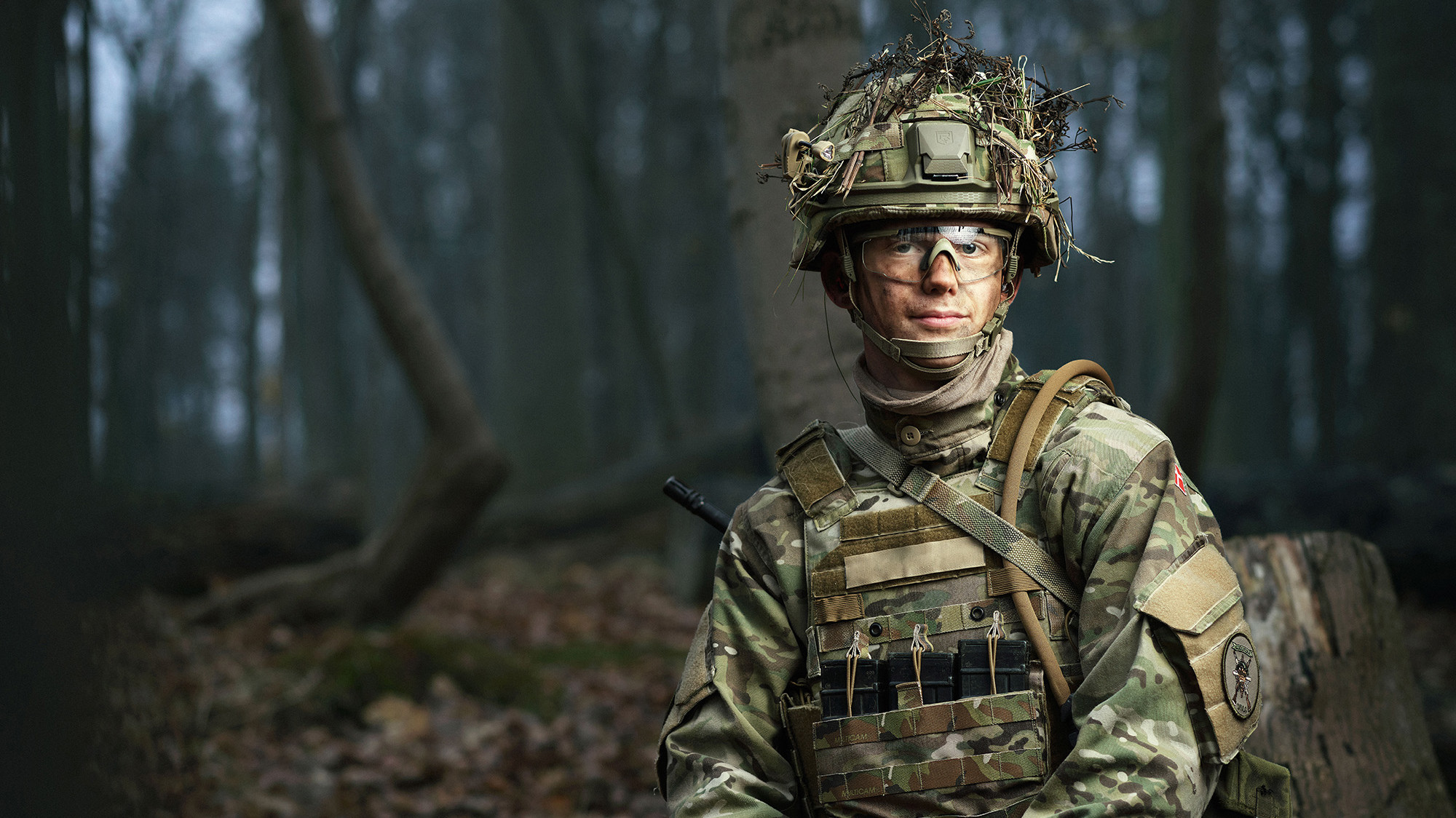 Soldat i camouflageuniform med hjelm pyntet med grene og beskyttelsesbriller står i skovmiljø med taktisk udstyr.