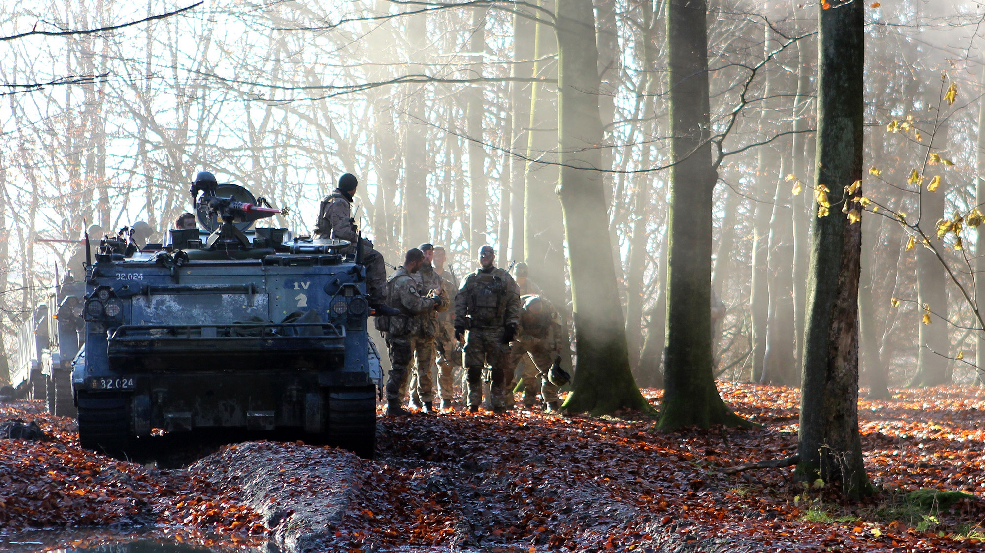 Soldater i camouflage uniformer taler sammen ved et slidt panserkøretøj i en tåget skov, omgivet af nedfaldne blade.