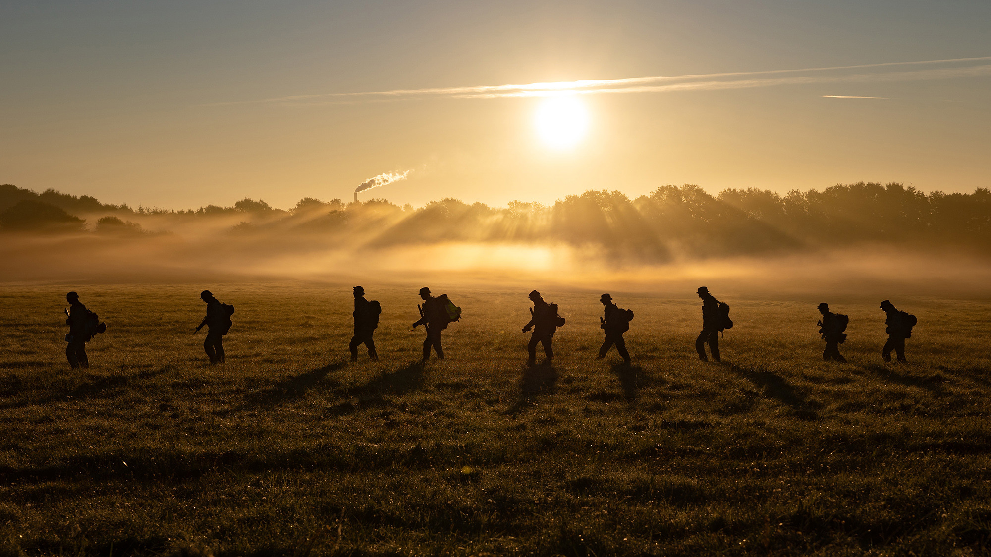 Silhuetter af personer med rygsække går gennem en tåget mark ved solopgang, varm sol belyser tågen og en skorstensrøg i baggrunden.