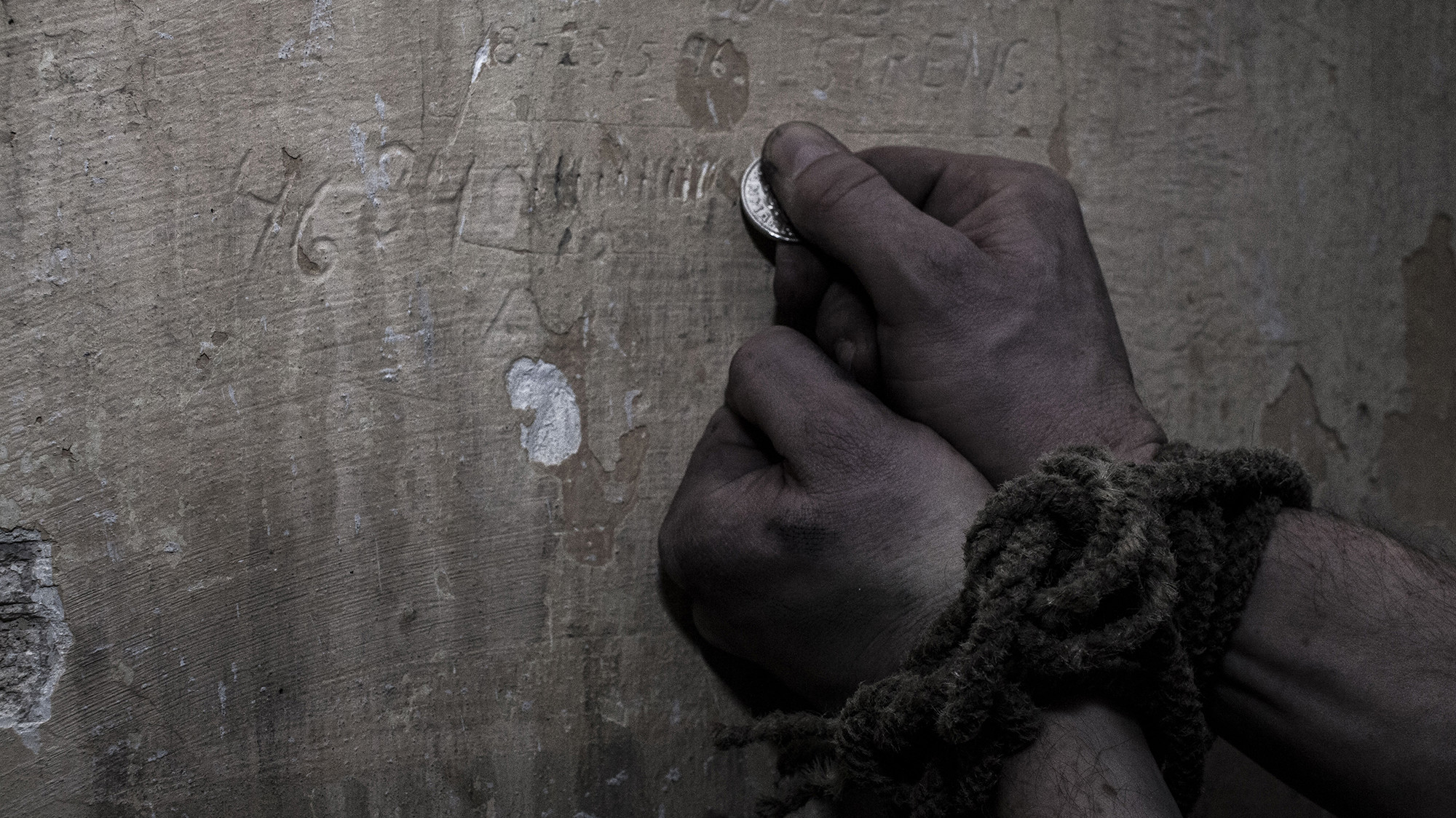 To hænder, den ene holder en lille mønt, med snavsede fingre og et håndled bundet med reb foran en slidt trævæg. Stemningen er dyster.