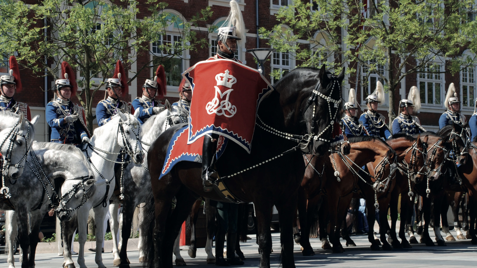 Rytter i rød og blå kappe med kroneemblem foran andre ceremonielle soldater til hest ved grønt område og murstensbygning.