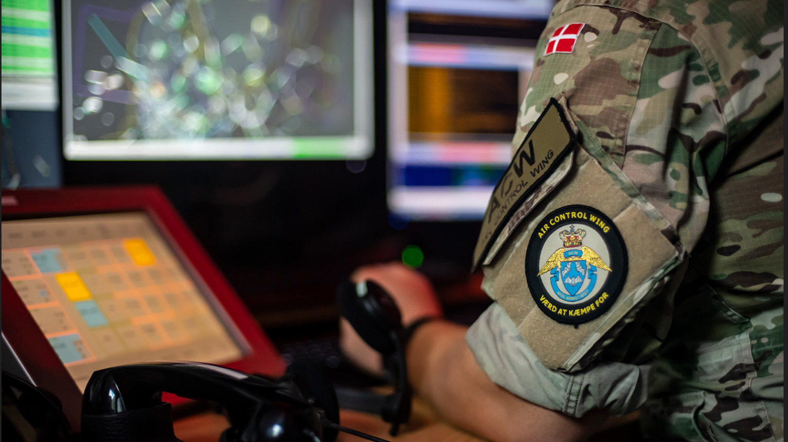 Soldat i camouflageuniform med "Air Control Wing"-mærke og dansk flag ved kontrolpanel og skærme med data i et operationsrum.
