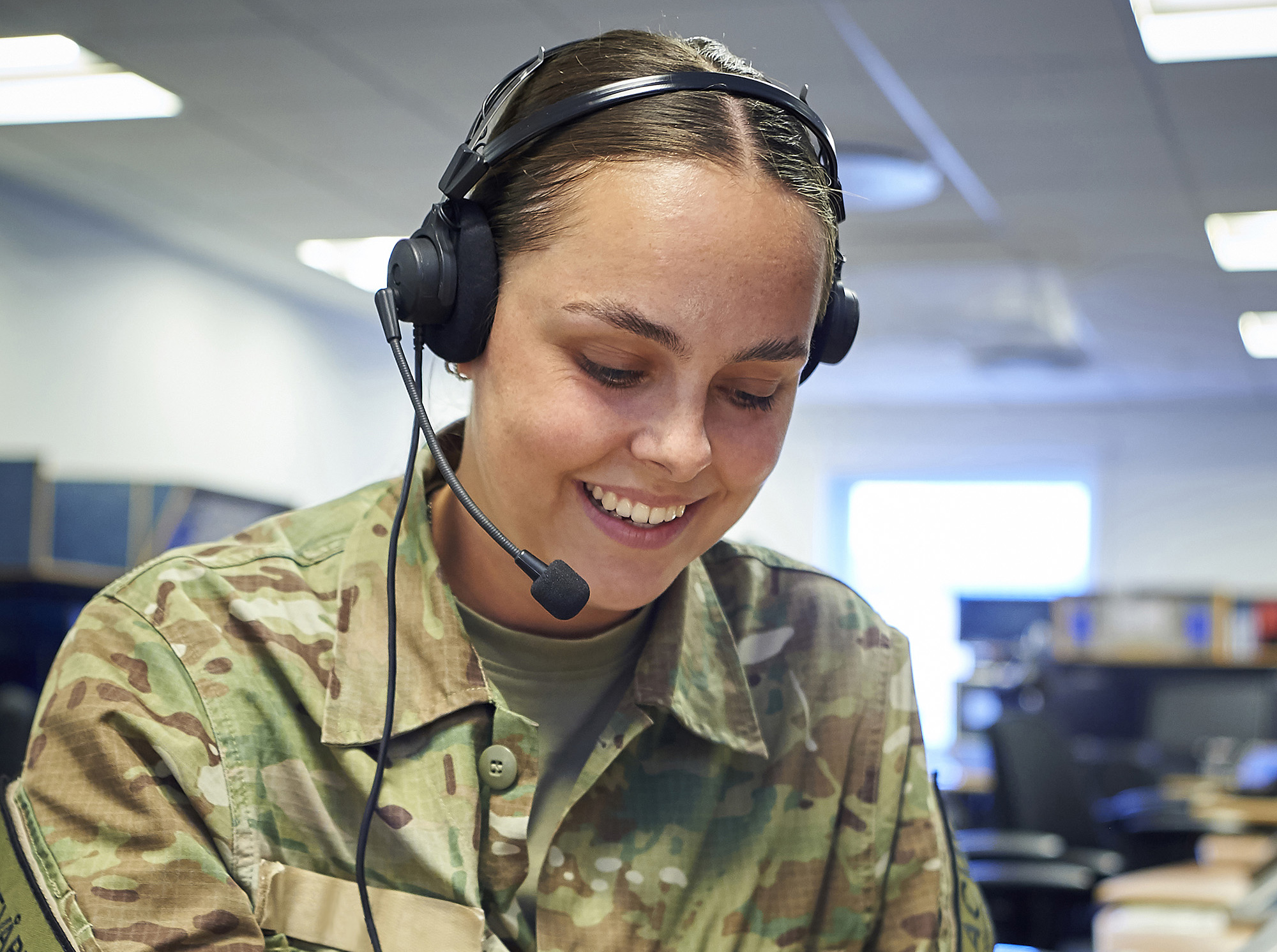 En ung kvinde iført militæruniform med camouflage og headset med mikrofon er hovedmotivet. Hun smiler, hvilket antyder en positiv interaktion, muligvis i en kommunikations- eller supportrolle. Baggrunden viser et moderne arbejdsområde, og hendes uniform bærer militære insignier. Hendes udtryk er opmærksomt og professionelt.