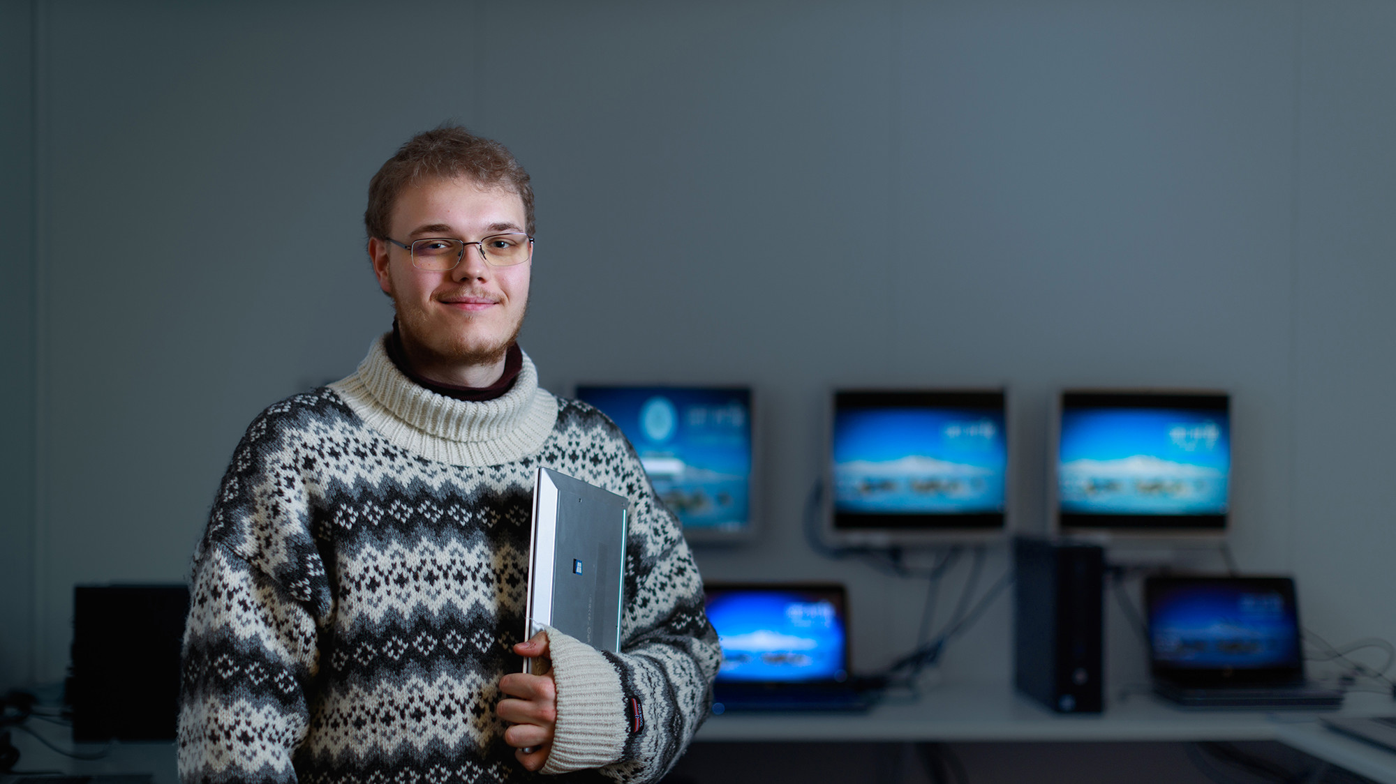 Ung mand med briller smiler og holder en tablet, iført mønstret sweater, foran computer-skærme i et teknologisk miljø.