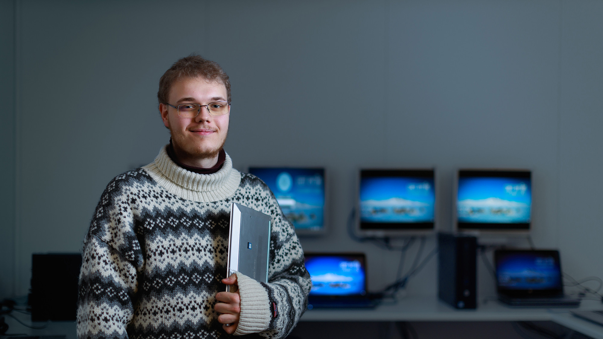 Ung mand med briller smiler og holder en bærbar under armen, iført mønstret sweater. Bag ham ses skærme med blå landskaber.