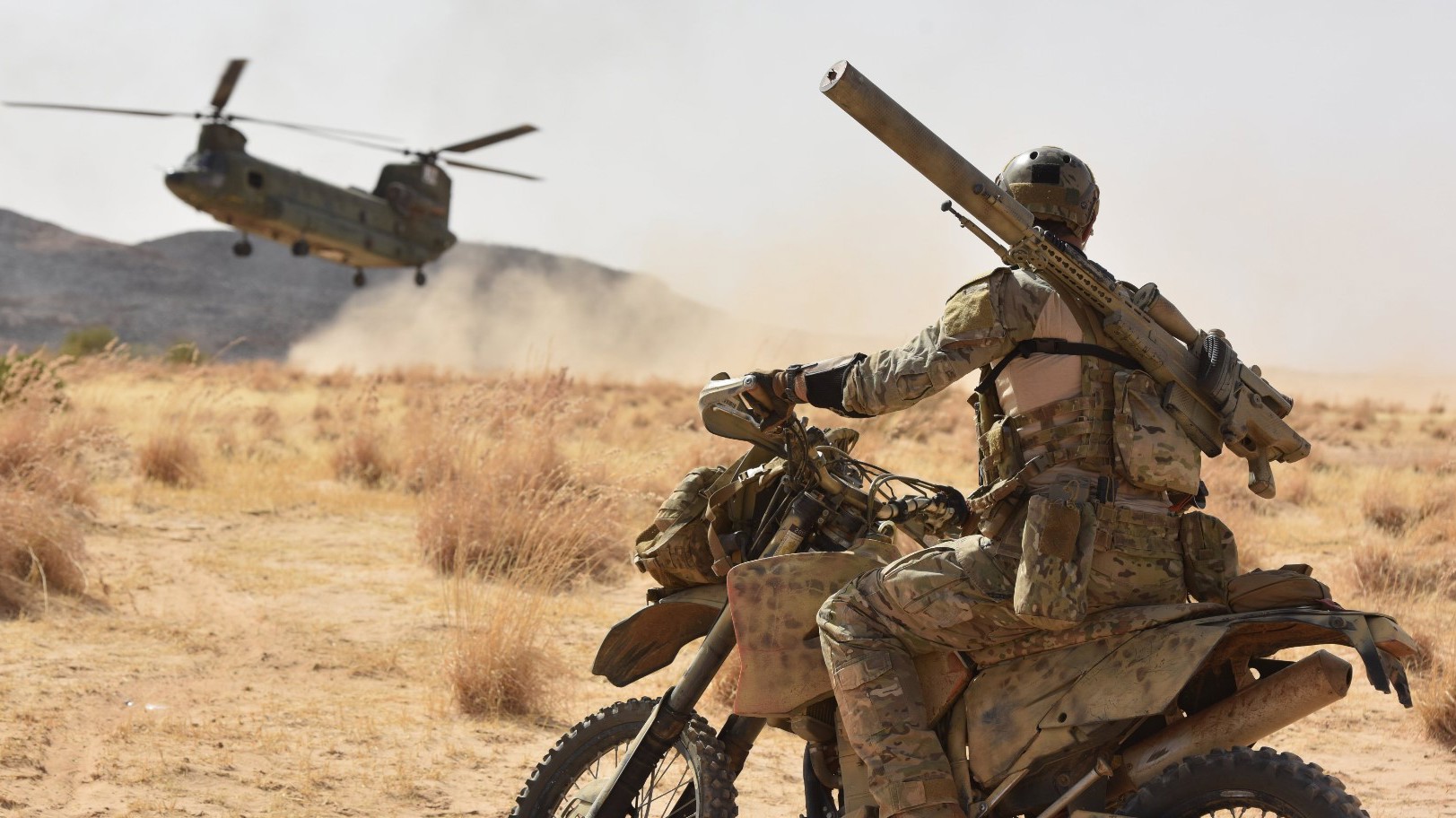 Soldat med taktisk udstyr og missilaffyrer på motorcykel, militærhelikopter svæver i støvet over ørkenagtigt landskab.