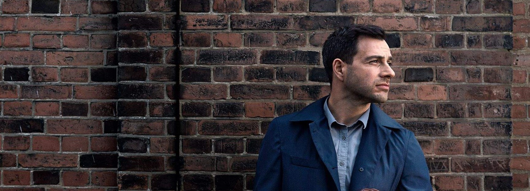 En mand i blå jakke og grå skjorte står foran en mur af røde mursten og kigger ud til højre med et eftertænksomt udtryk.