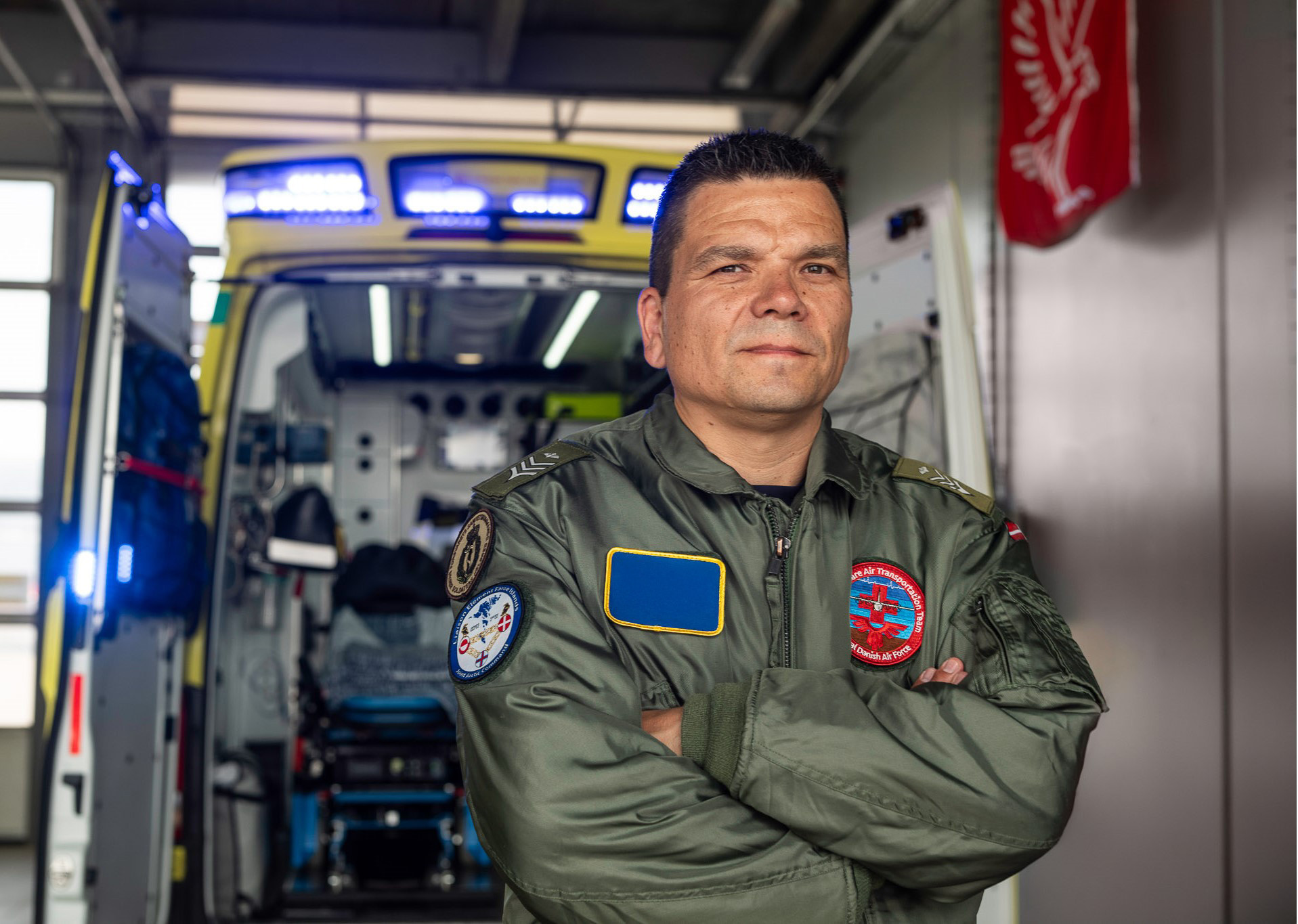 Militærperson i grøn flyverdragt står med krydsede arme foran en ambulance med åben bagdør.