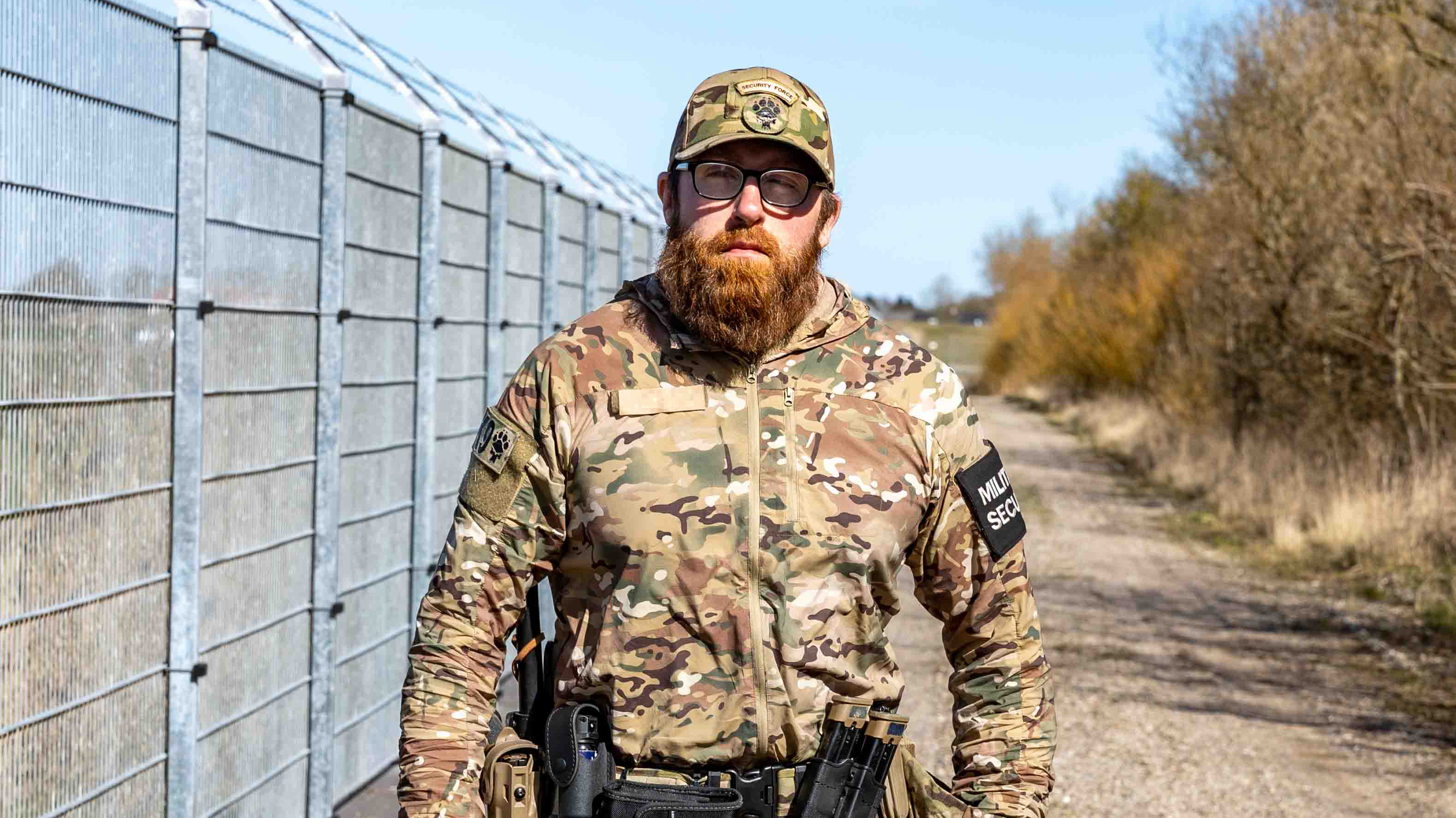 Skægget person i camouflagejakke og kasket står selvsikkert foran højt hegn med taktisk udstyr og værktøj på bæltet, omgivet af træer og blå himmel.