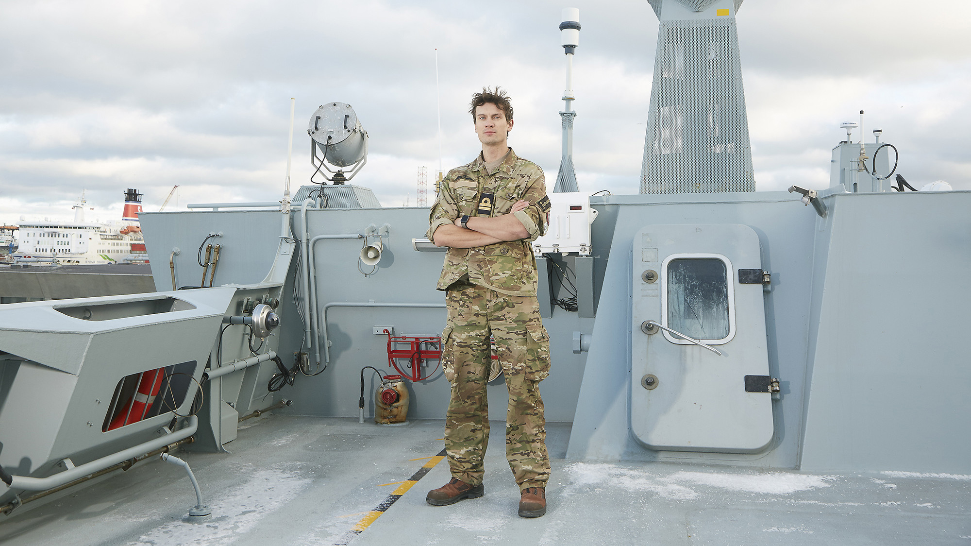 En soldat står på dækket af et marineskib iført camouflageuniform med armene over kors og et selvsikkert udtryk. Baggrunden viser forskelligt marineudstyr og strukturer, der indikerer skibets operative miljø. I det fjerne ses et civilt fartøj, hvilket antyder, at skibet ligger til kaj eller er nær havn. Scenen udstråler beredskab og professionalisme i et maritimt miljø.