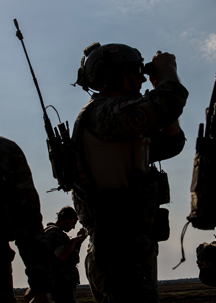 Silhuetter af soldater i taktisk udstyr og hjelme, en med kikkert, foran dramatisk, skyet himmel.