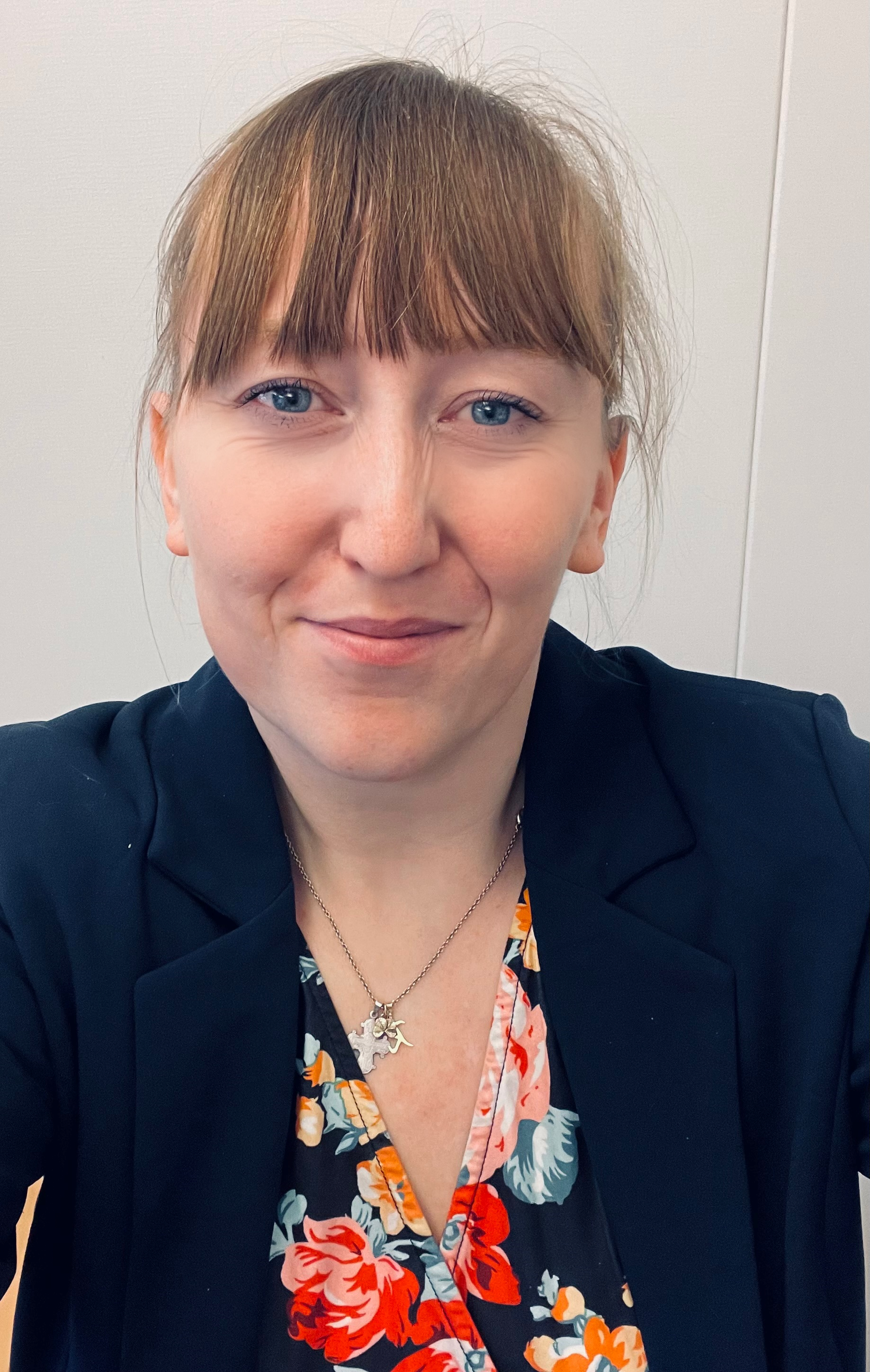 Billedet viser en kvinde med lysebrunt hår med pandehår og blå øjne. Hun har en mørk blazer på over en blomstret top i livlige farver, og hun bærer en sølvhalskæde med et vedhæng. Baggrunden er ensfarvet og neutral, hvilket fremhæver hendes venlige og imødekommende udtryk.