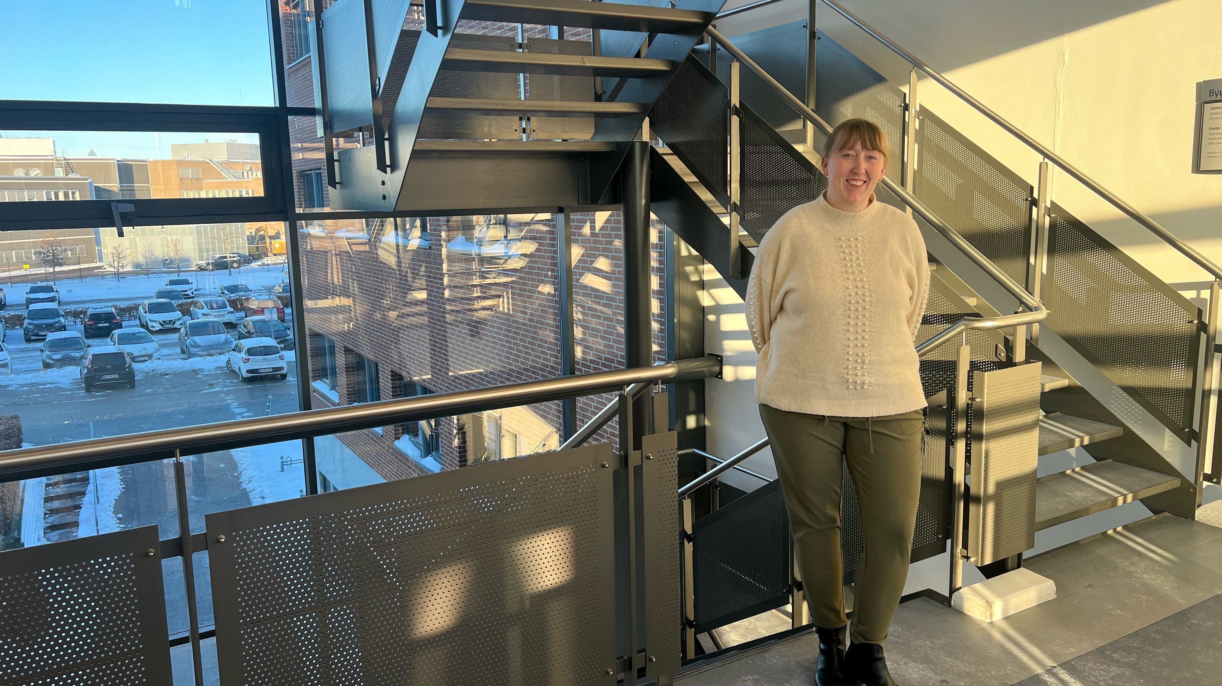 En person står ved siden af en moderne trappe i en lyst, nutidigt byggeri. Personen har en beige sweater med struktureret mønster og olivengrønne bukser på. I baggrunden ses store vinduer med udsigt til en parkeringsplads med flere biler og et snedækket landskab, hvilket antyder et koldt klima. Stemningen er lys og indbydende med fokus på bygningens moderne arkitektur.