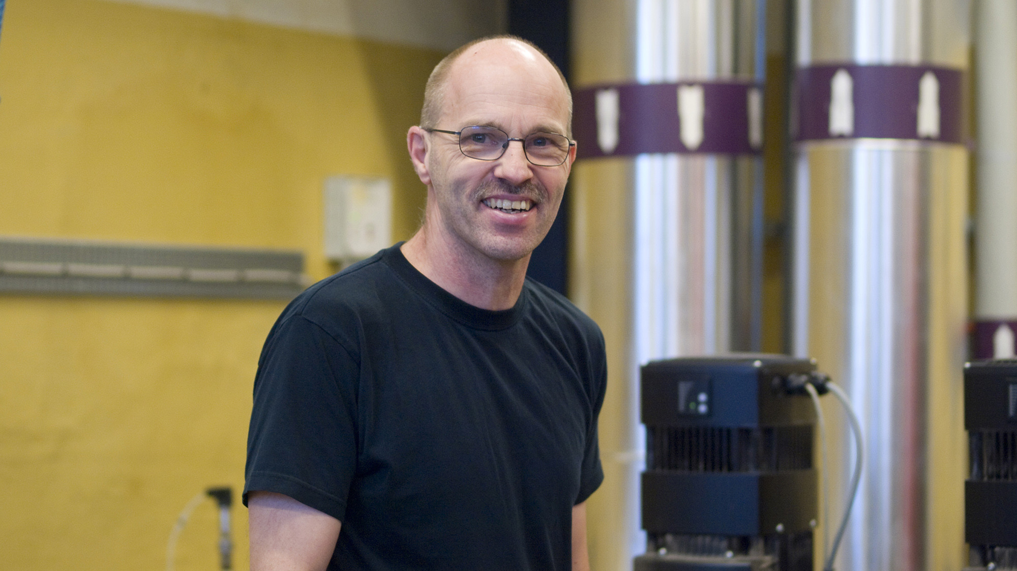 Mand med venligt udtryk og briller står i et teknisk værksted med metallisk udstyr og cylindriske genstande i baggrunden.