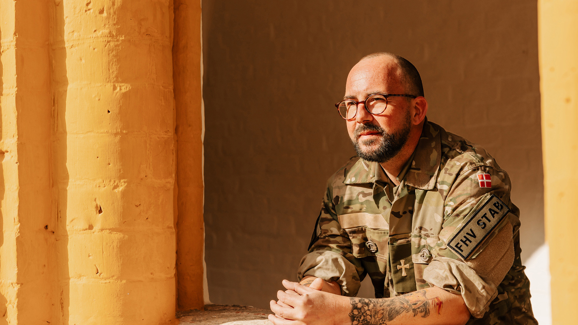 Mand i camouflageuniform med enheds-patch, briller og tatoveringer på underarmene sidder tænksomt foran gule søjler.