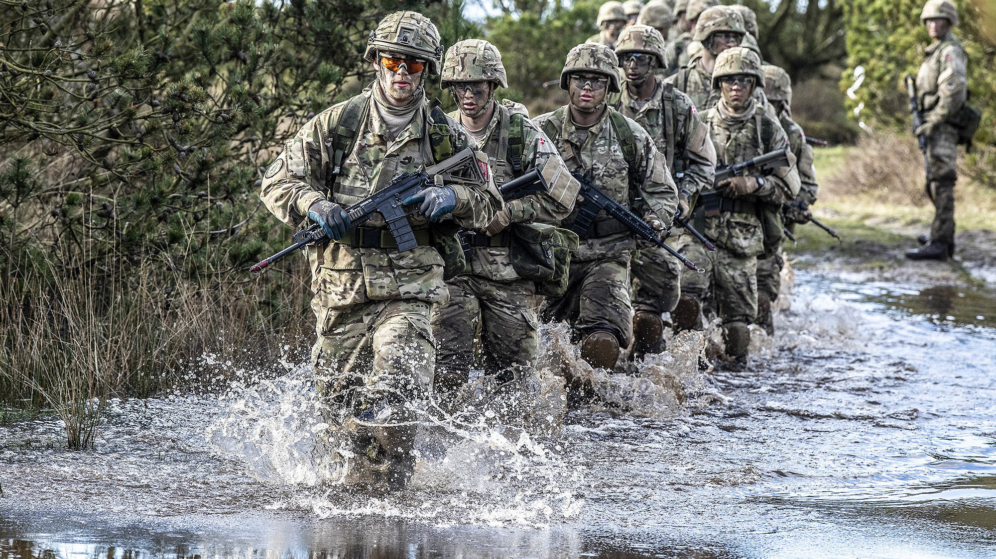 Soldater i camouflageuniformer krydser lavt vand under øvelse, ledes af en bevæbnet soldat med beskyttelsesbriller.