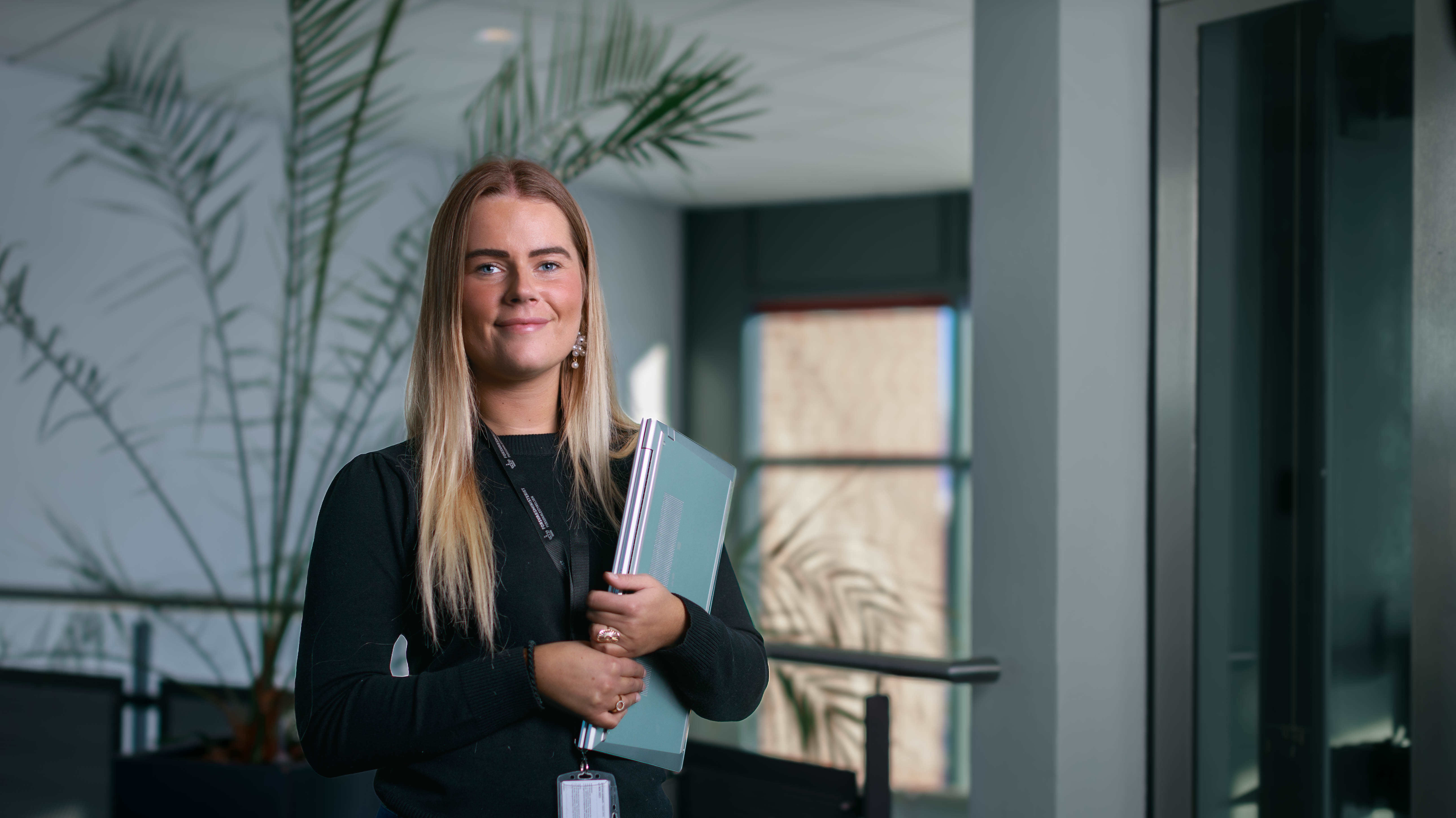 Ung kvinde med mapper i hånden, iført sort bluse, står i et lyst, moderne lokale med store vinduer og grønne planter.